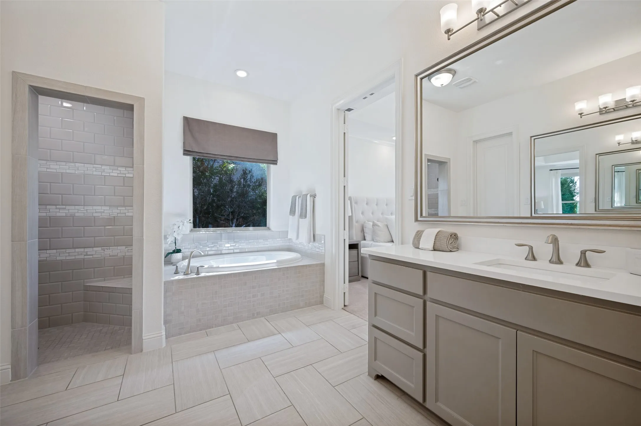 Tile-surround soaking tub with a window, walk-in shower with bench seating, single vanity with a white countertop, large wall-mounted mirror, and tile flooring