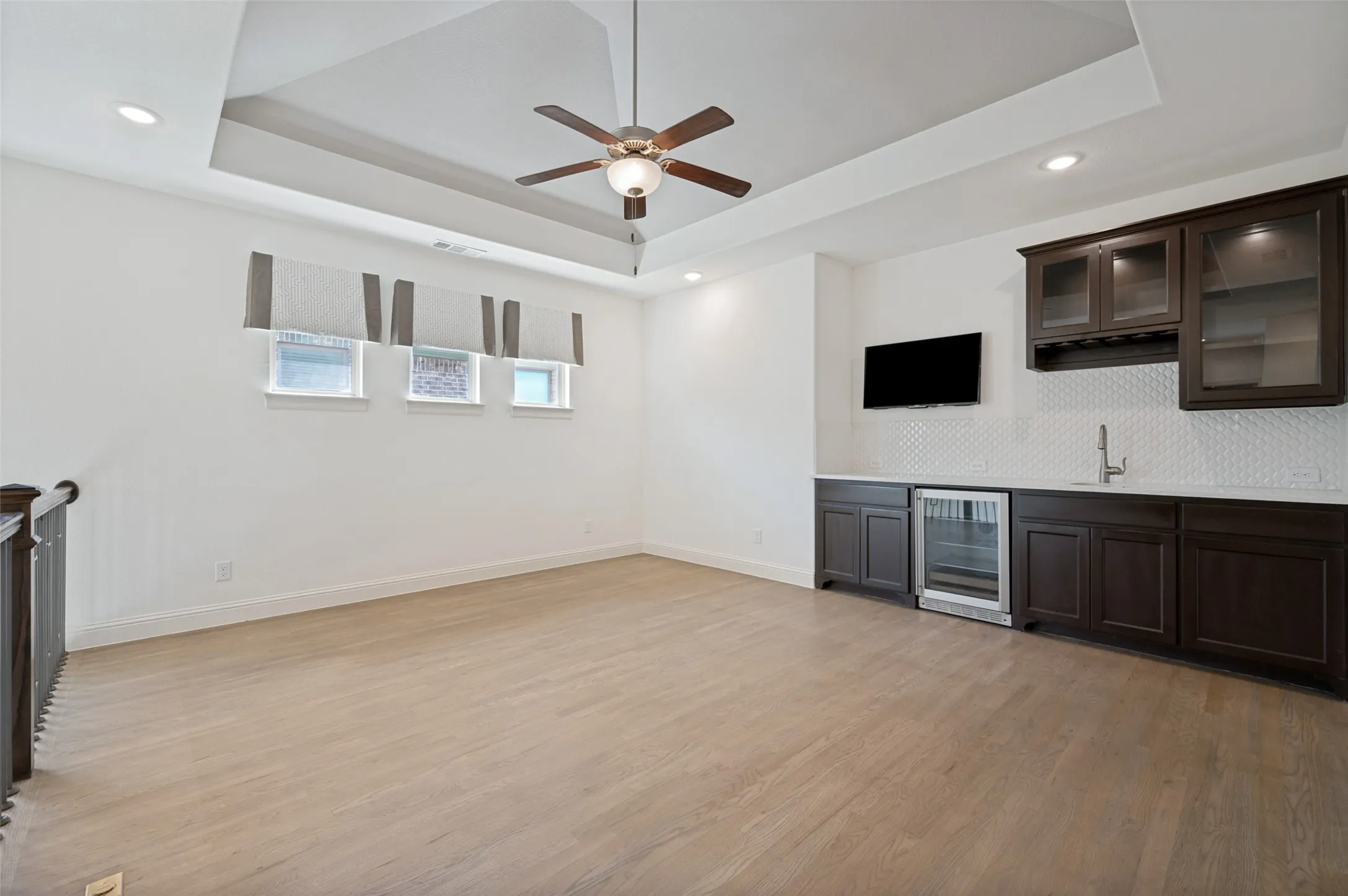 Recessed lighting, wood-finish flooring, tray ceiling with a ceiling fan, and built-in wet bar with a beverage cooler
