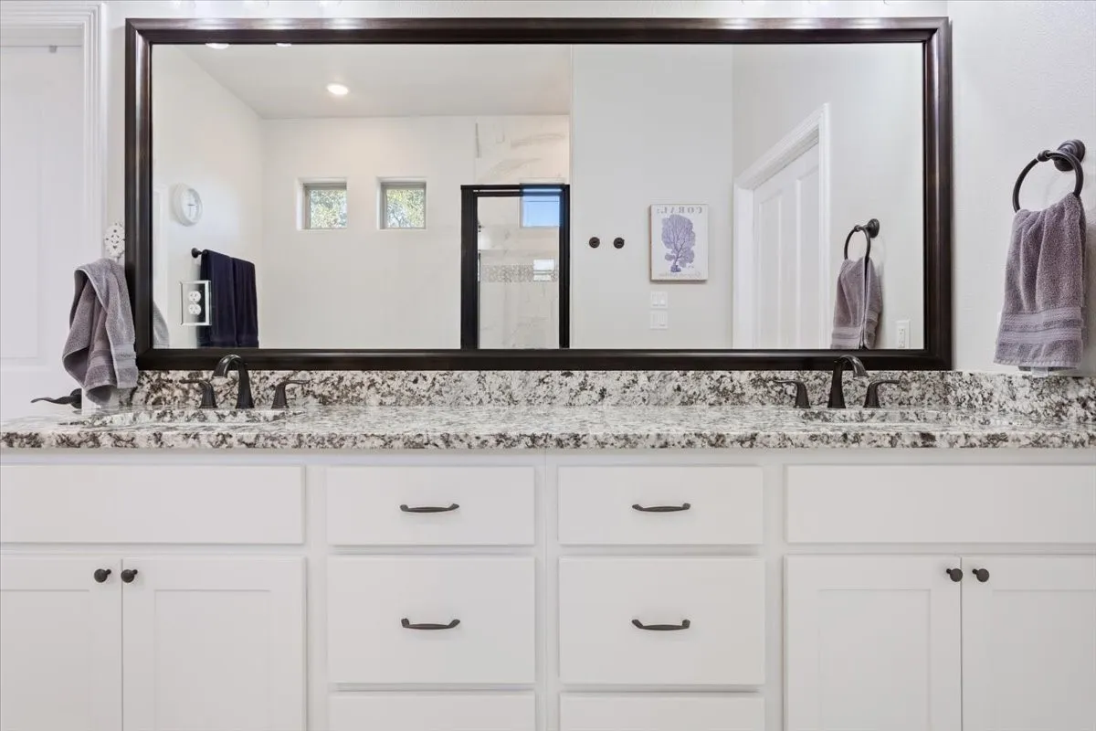 Bathroom vanity featuring a speckled countertop, dual sinks with dark fixtures, and white cabinetry with dark hardware