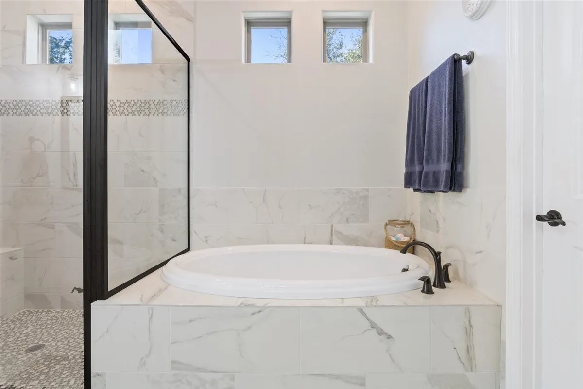 The bathroom features a built-in bathtub with a dark bronze faucet, a walk-in shower with a glass enclosure and pebble tile flooring, and white tiled walls with a decorative accent band