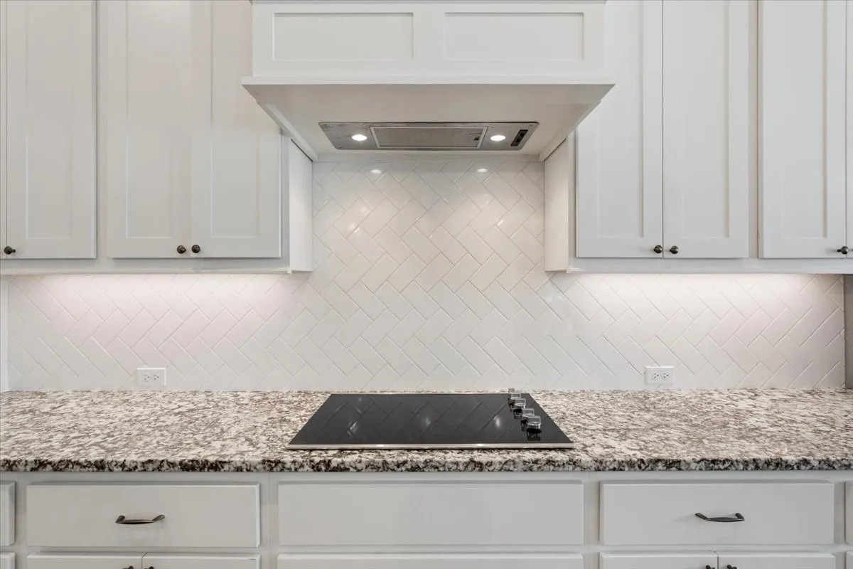 The kitchen features white cabinetry, granite countertops, a herringbone tile backsplash, and an integrated cooktop with an overhead range hood