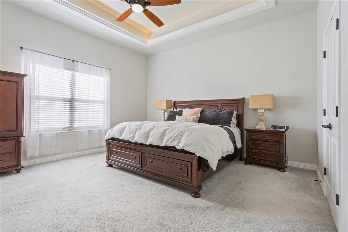 Bedroom featuring light gray carpeting, a large window with blinds and sheer curtains, and a tray ceiling with a ceiling fan