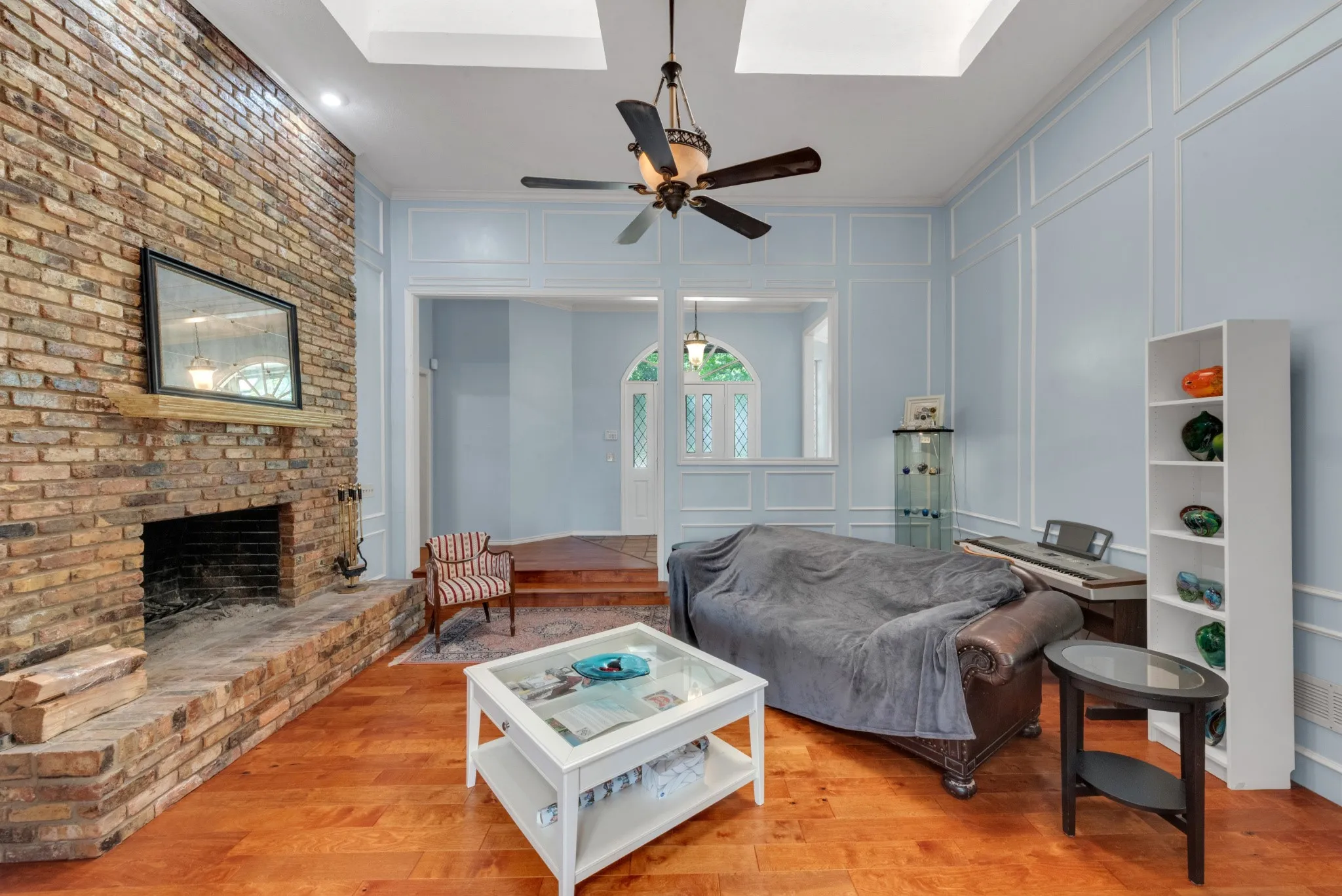 Living area featuring hardwood floors, a prominent brick fireplace with a wood mantle, and light blue walls with decorative paneling