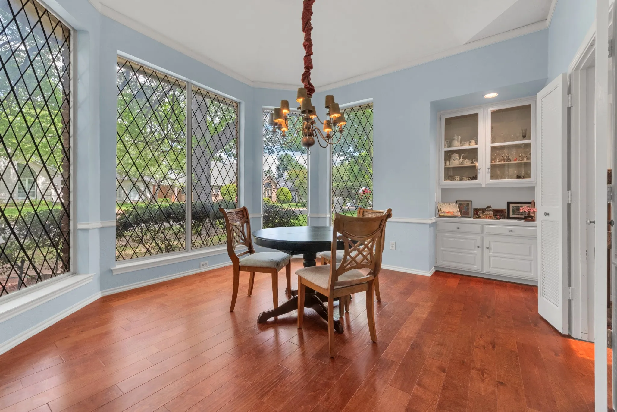 This space features abundant natural light from the bay windows with decorative grilles, hardwood flooring, and a built-in cabinet with glass-fronted upper doors
