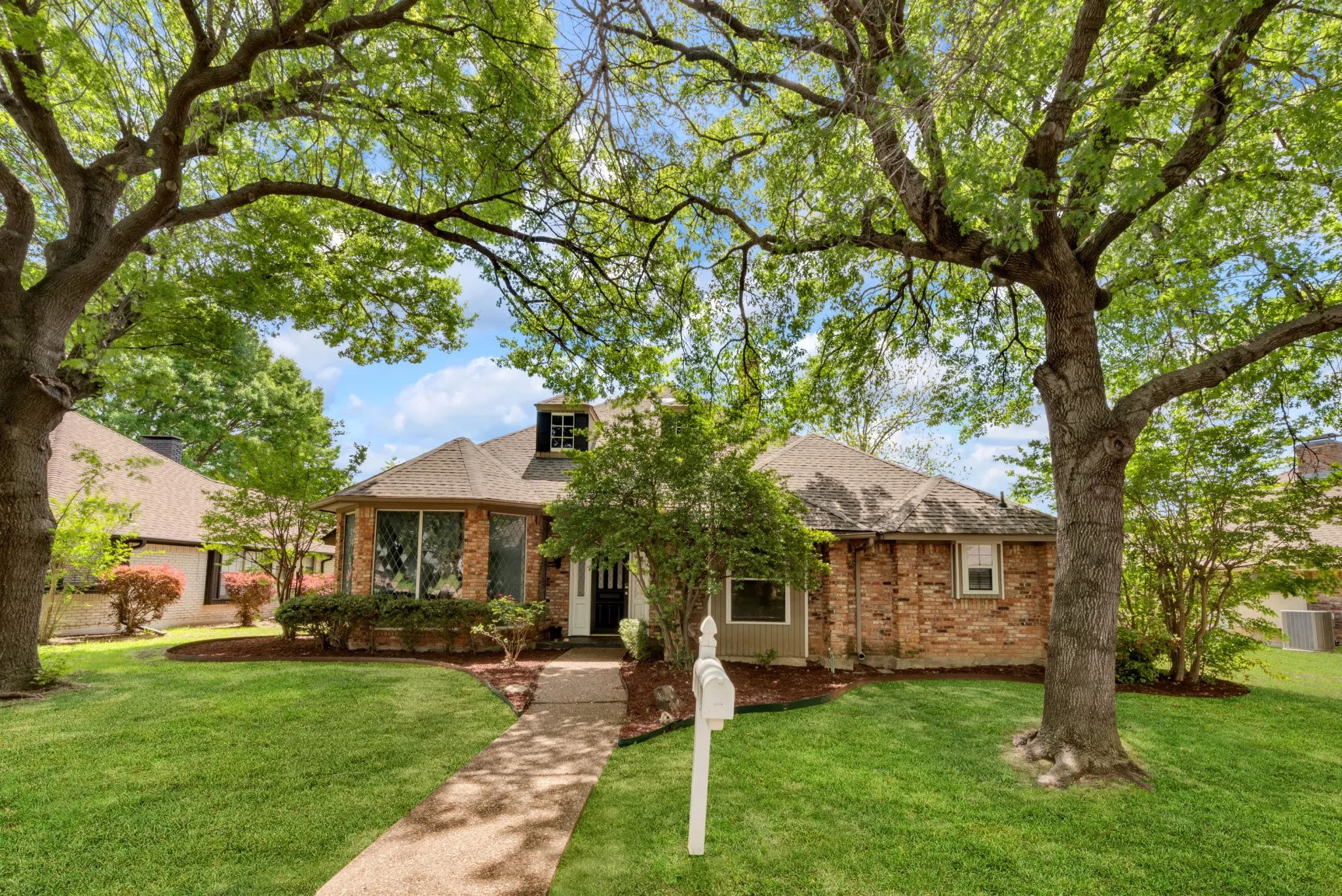 The property features a brick exterior, a well-maintained lawn, and mature trees providing natural shade