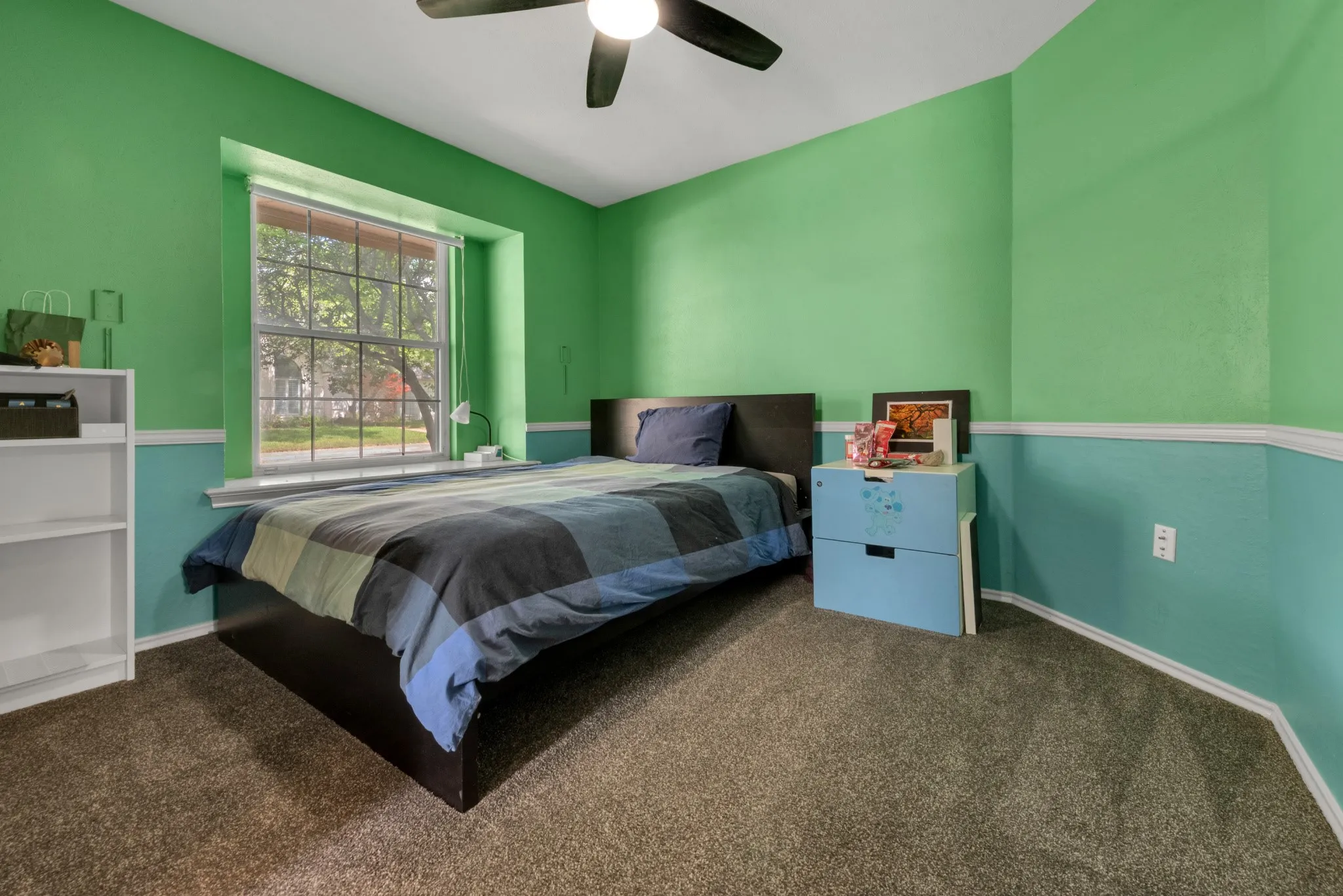 The room features carpeted flooring, a window with a view of greenery, and a ceiling fan with integrated lighting