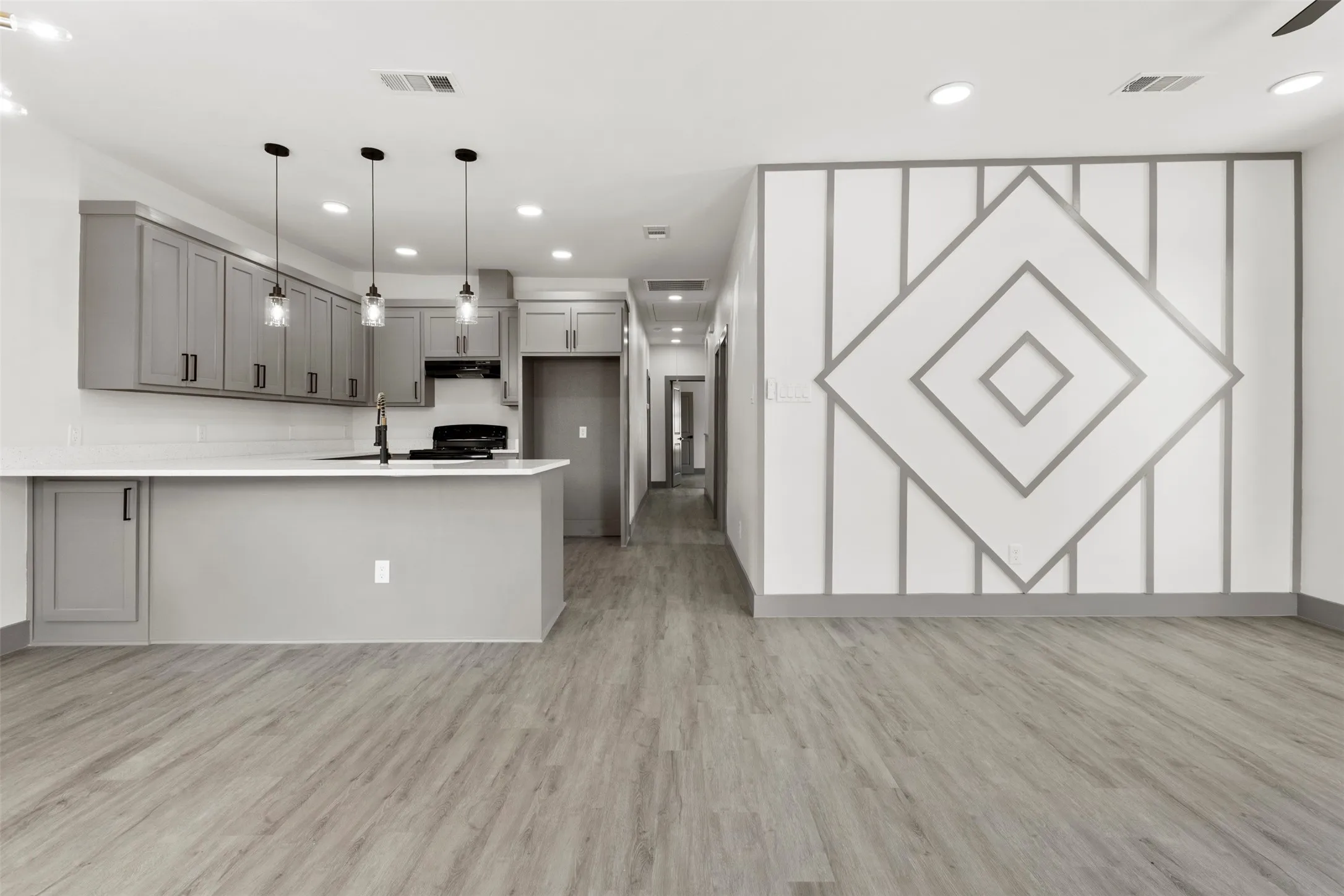 Open concept living area with light-colored flooring, a kitchen featuring grey cabinetry and pendant lighting, and a decorative accent wall