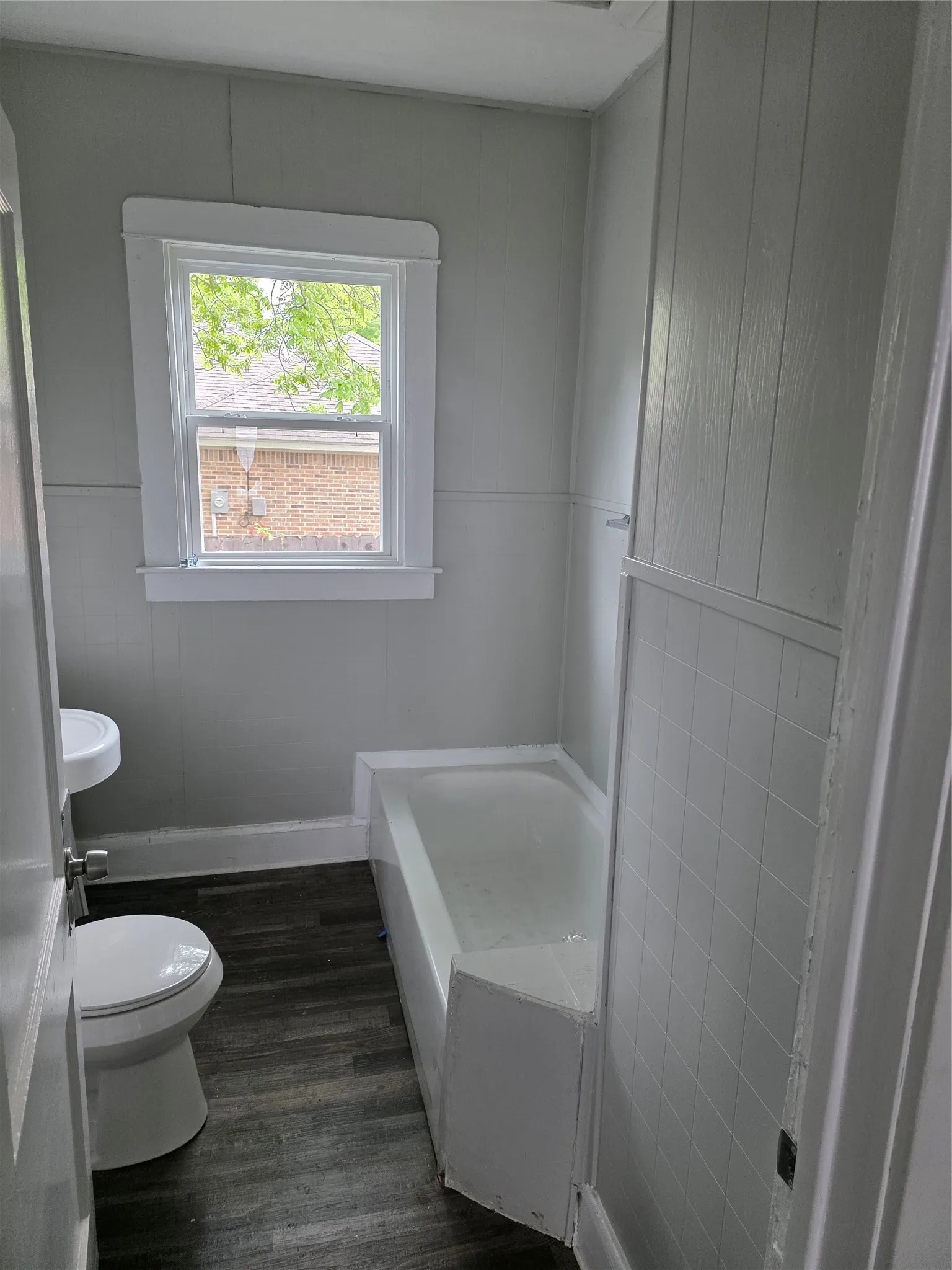 Bathroom featuring a window providing natural light, a bathtub, a toilet, a pedestal sink, and wood-look flooring