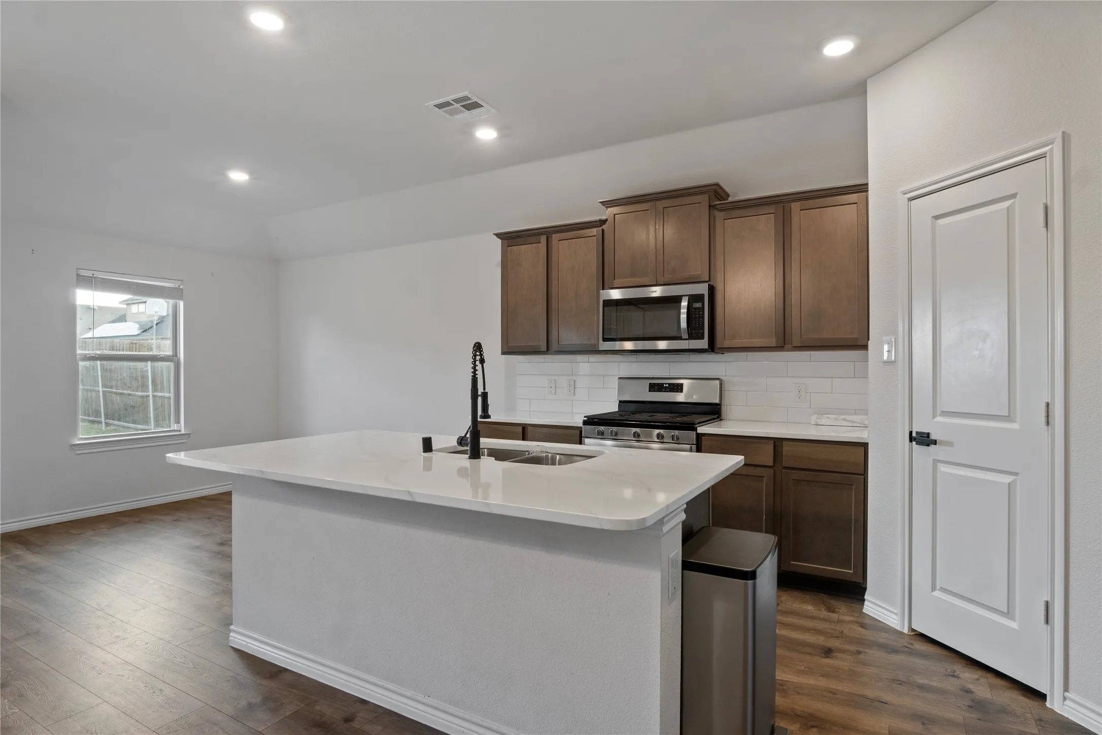 The kitchen features a large island with a countertop and a sink, stainless steel appliances including a microwave and range, wood cabinetry, and a white subway tile backsplash