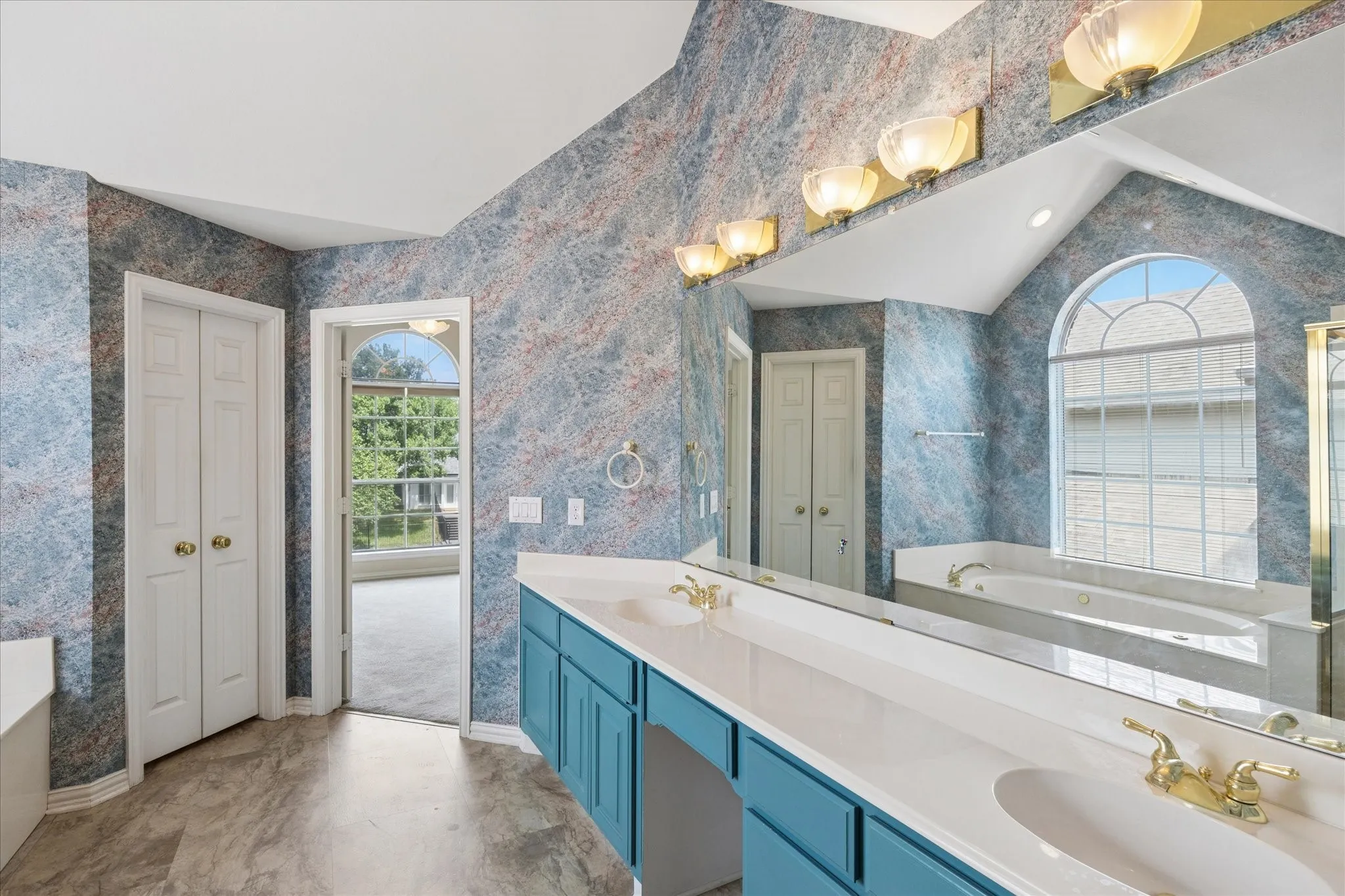 Bathroom featuring a double vanity with teal cabinetry and gold-toned fixtures, a large mirror, and an arched window above a bathtub