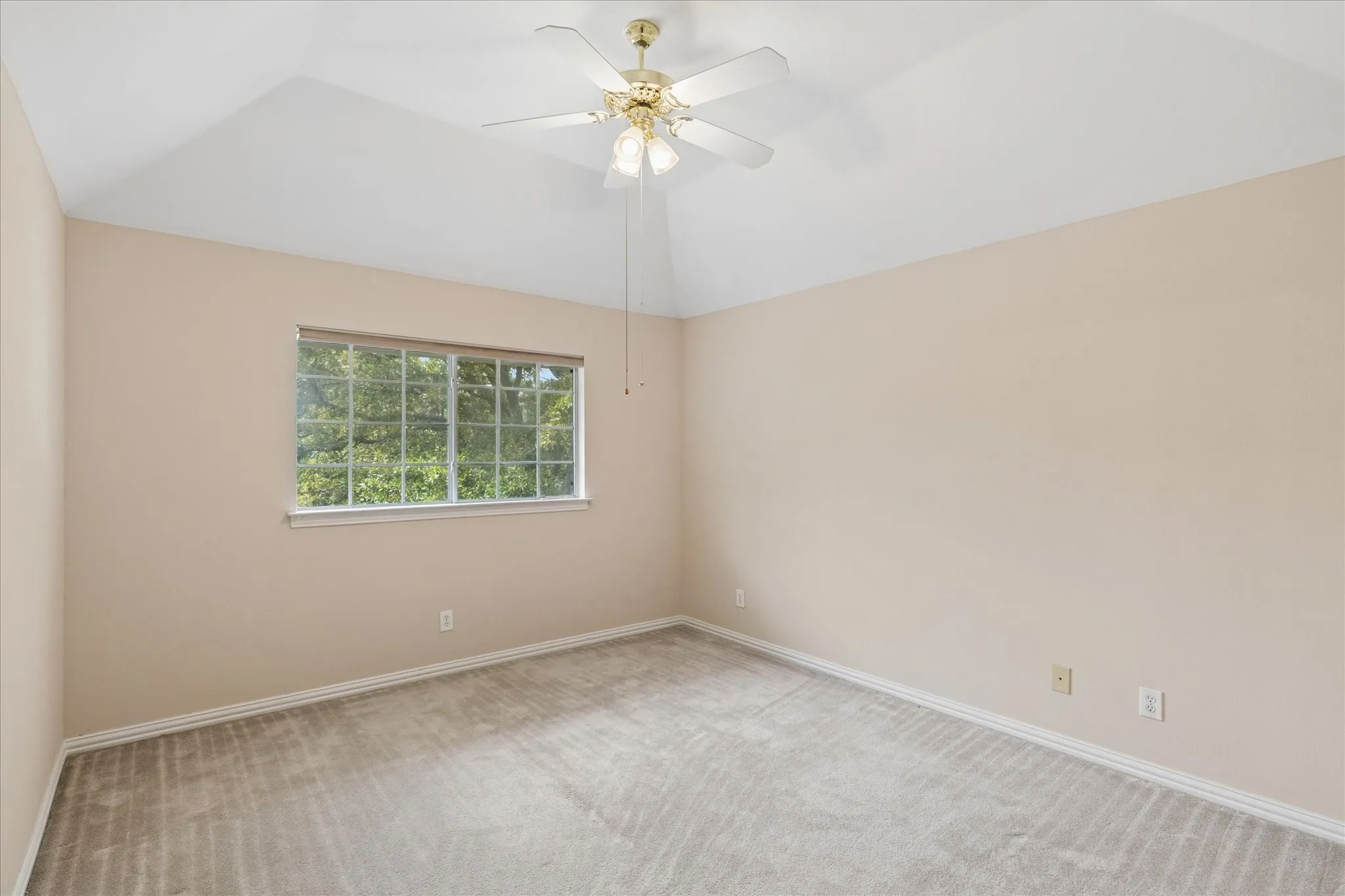 Inviting room featuring a vaulted ceiling with a ceiling fan, a window providing a view of green foliage, and light-colored carpet