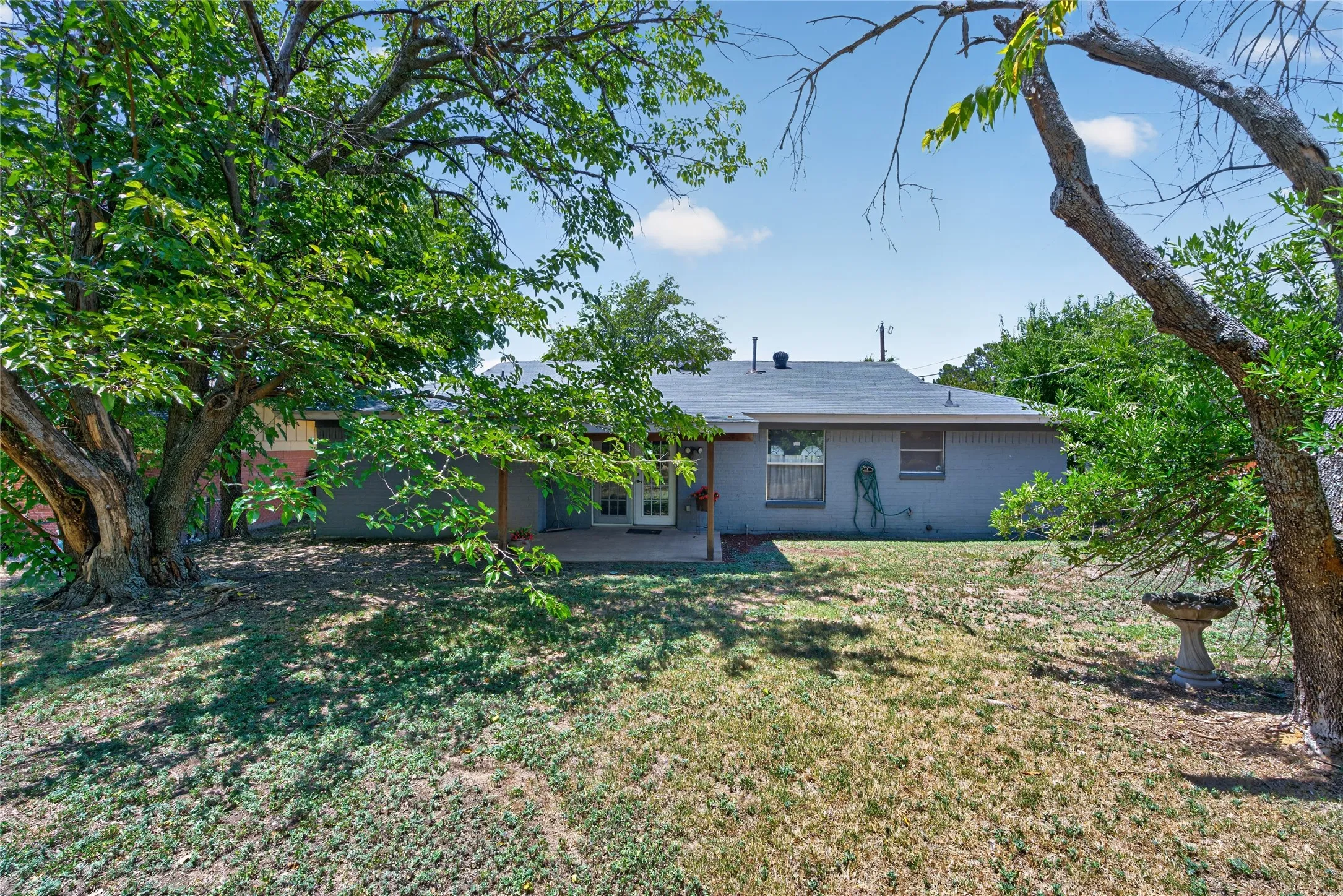 Spacious backyard featuring mature trees, a covered patio area, and a gray-painted exterior with multiple windows