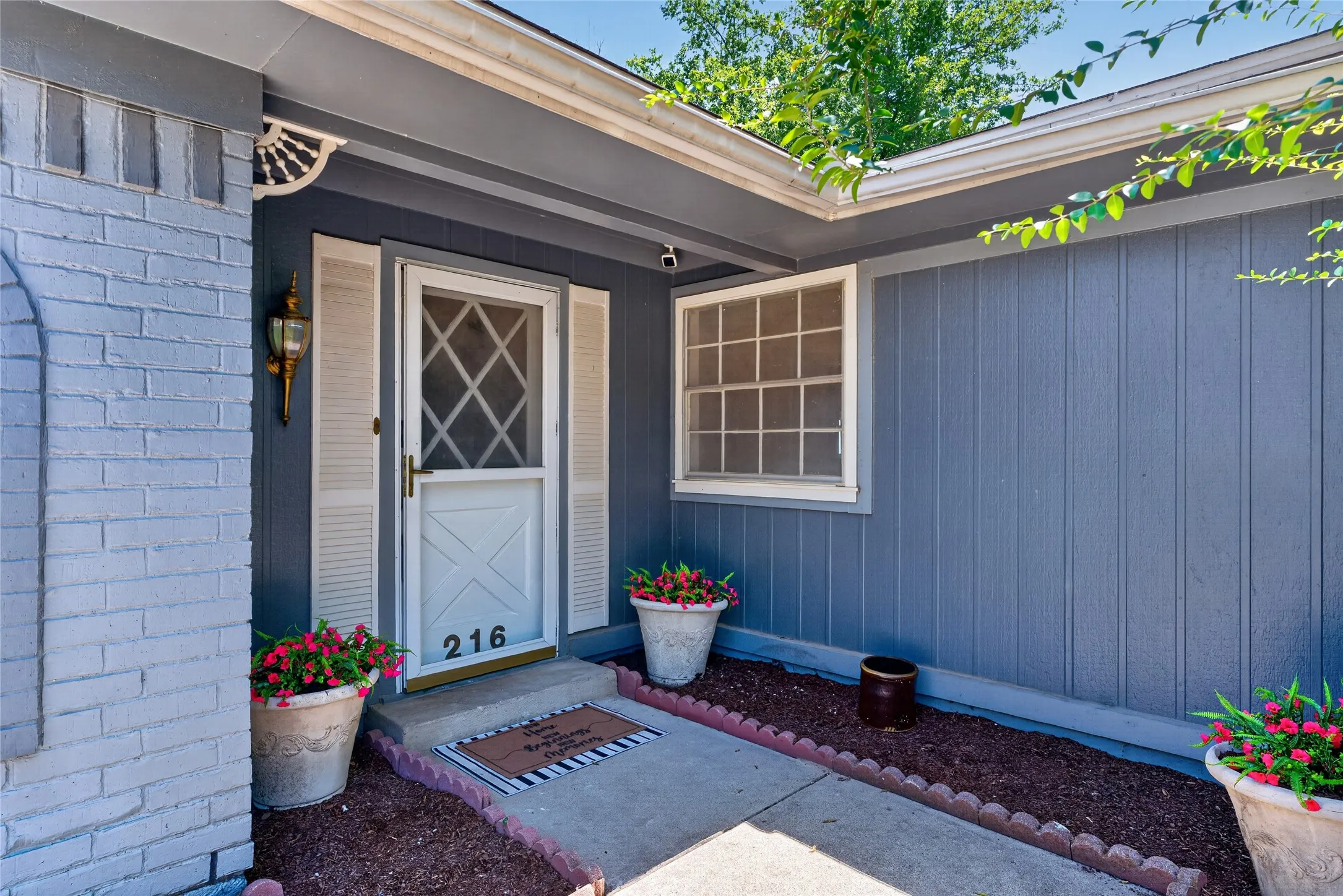 Entryway featuring a white storm door with geometric grille, white louvered shutters, and a brass exterior light fixture