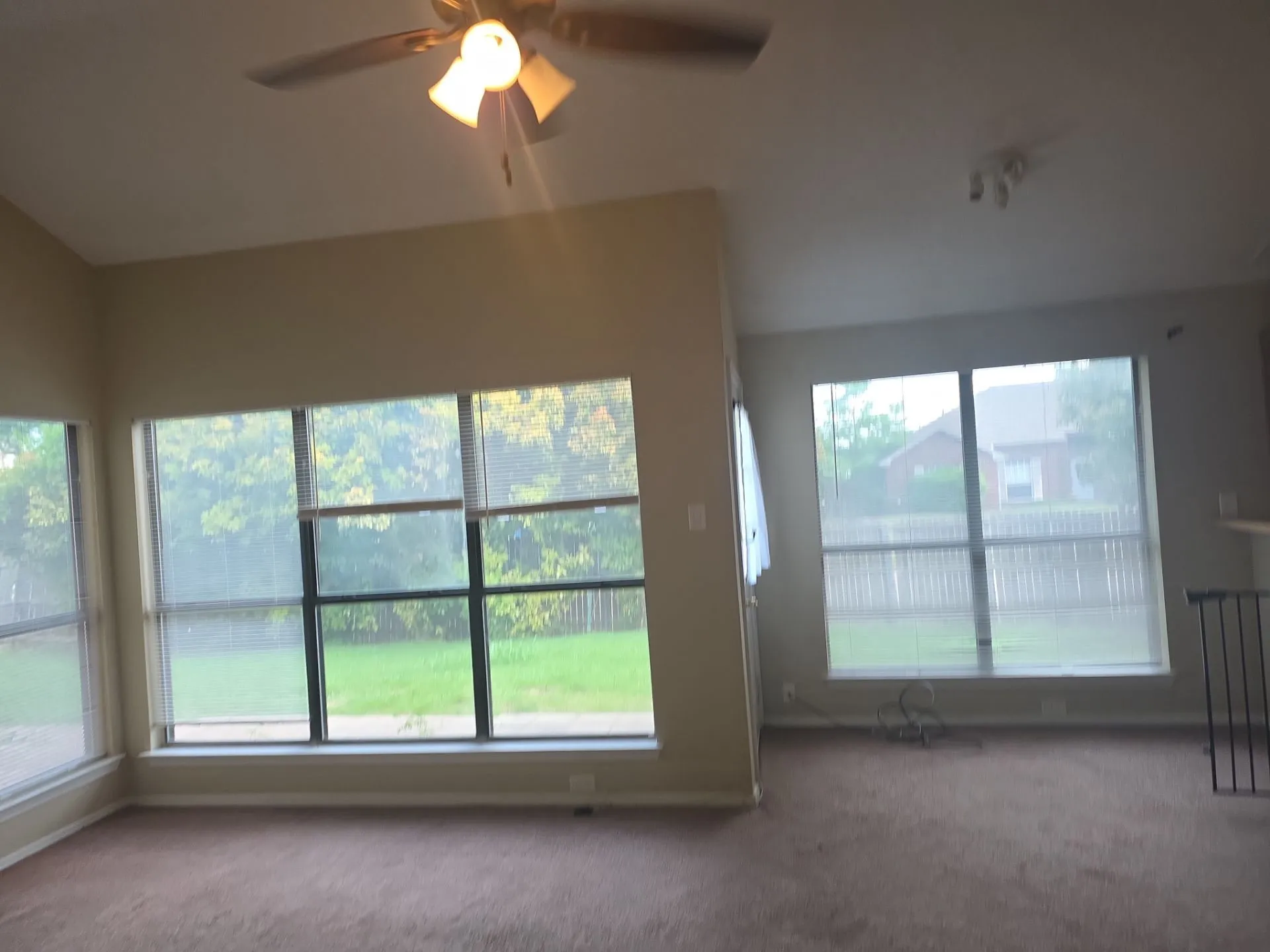 Spacious living area featuring carpeted flooring, large windows overlooking the yard, and a ceiling fan with light fixture