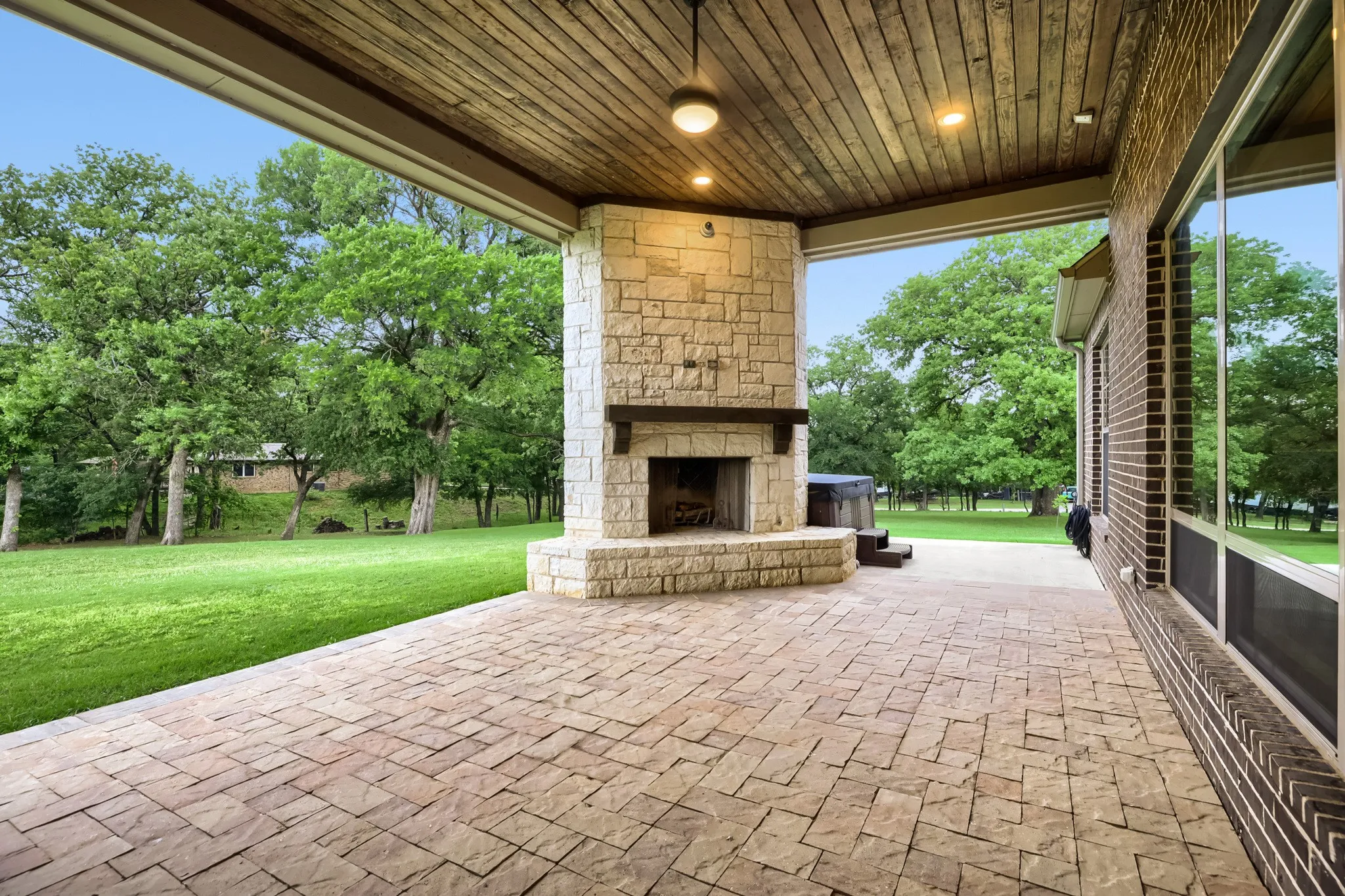 Covered outdoor patio featuring a stone-clad fireplace with a wood mantle, a wood-paneled ceiling with recessed lighting, and a brick-clad exterior wall with large windows