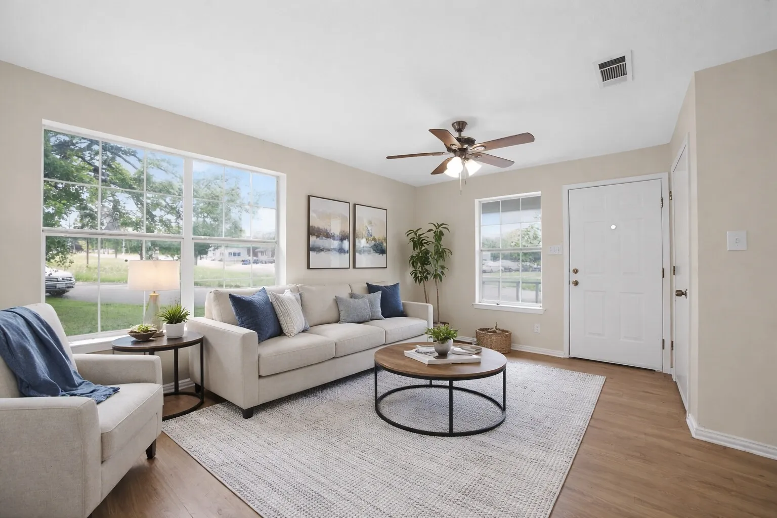 The living room features large windows, a ceiling fan with lighting, wood-style flooring, and a light-colored entry door - Virtually Staged