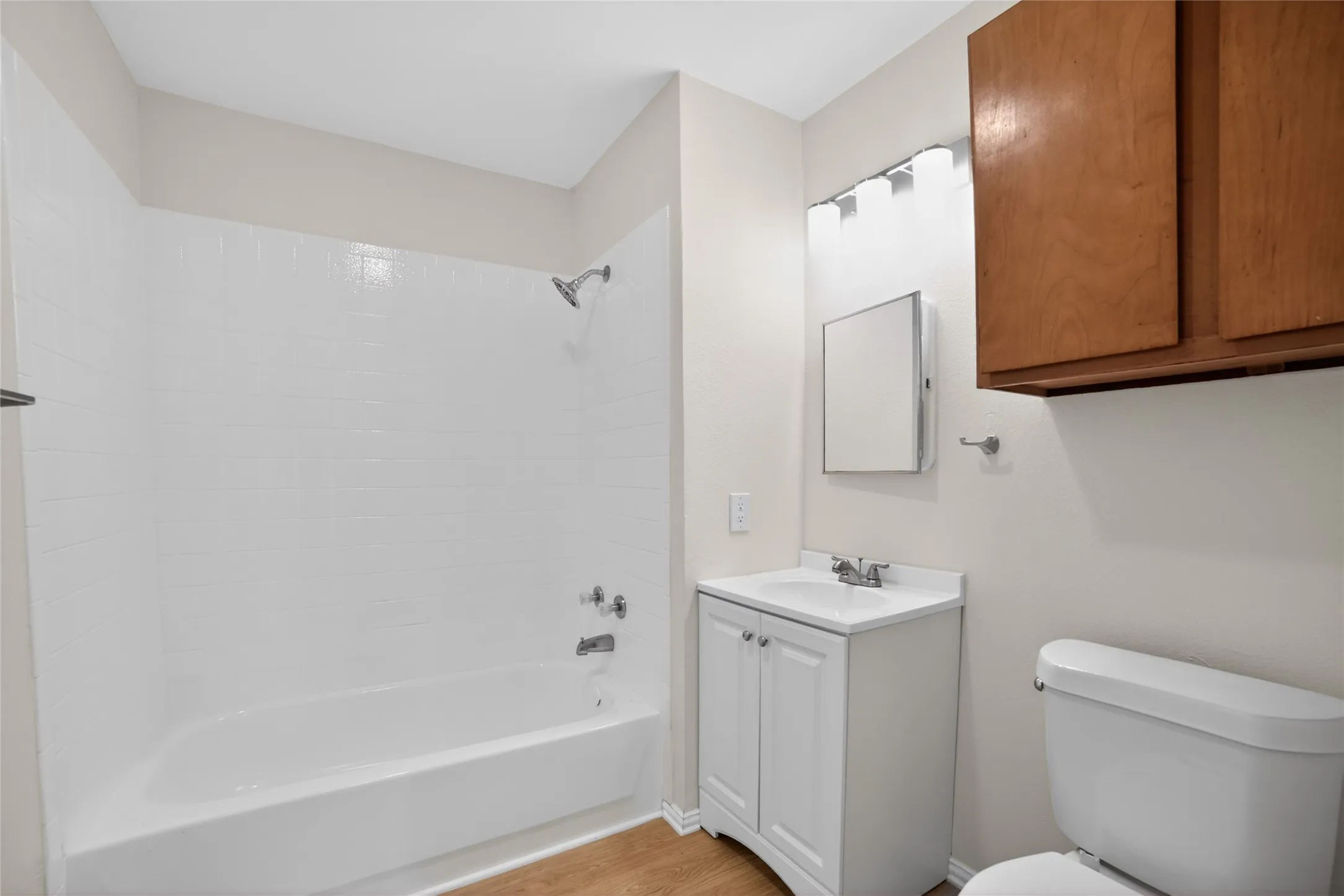 This bathroom features a shower and bathtub combination with white tiled walls, a white vanity with a sink, a wall-mounted mirror, and a toilet