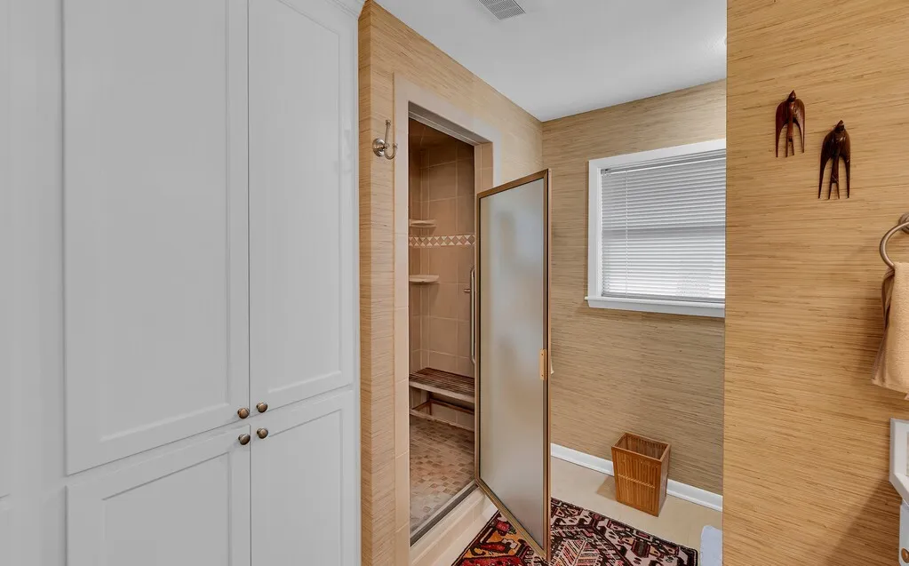 This bathroom features extensive built-in cabinetry, a glass-enclosed shower with tiled walls and a built-in bench, and a window with blinds