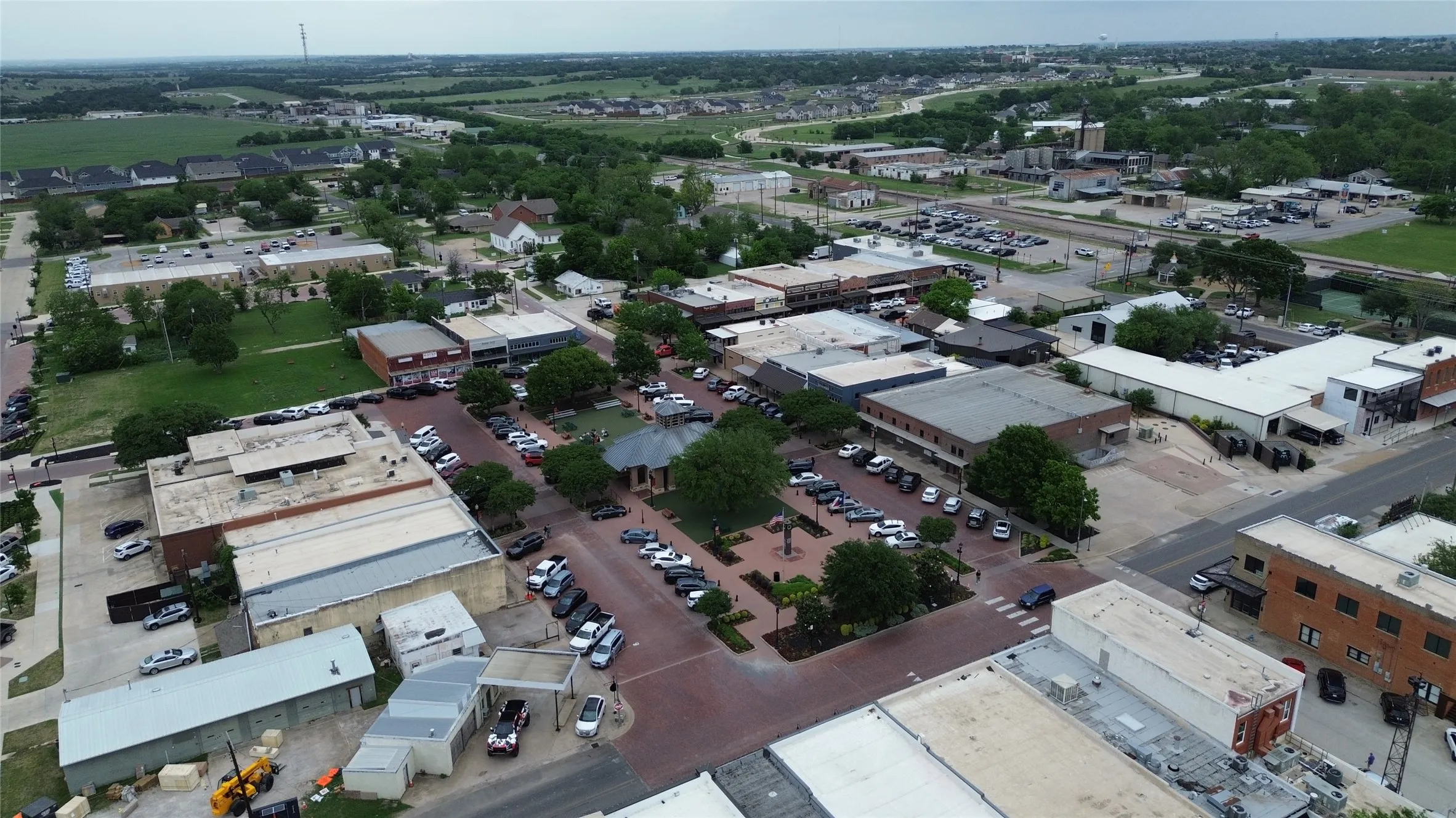Aerial view showcasing a community with various buildings, paved roads, and green spaces, including a central park area with trees and a gazebo