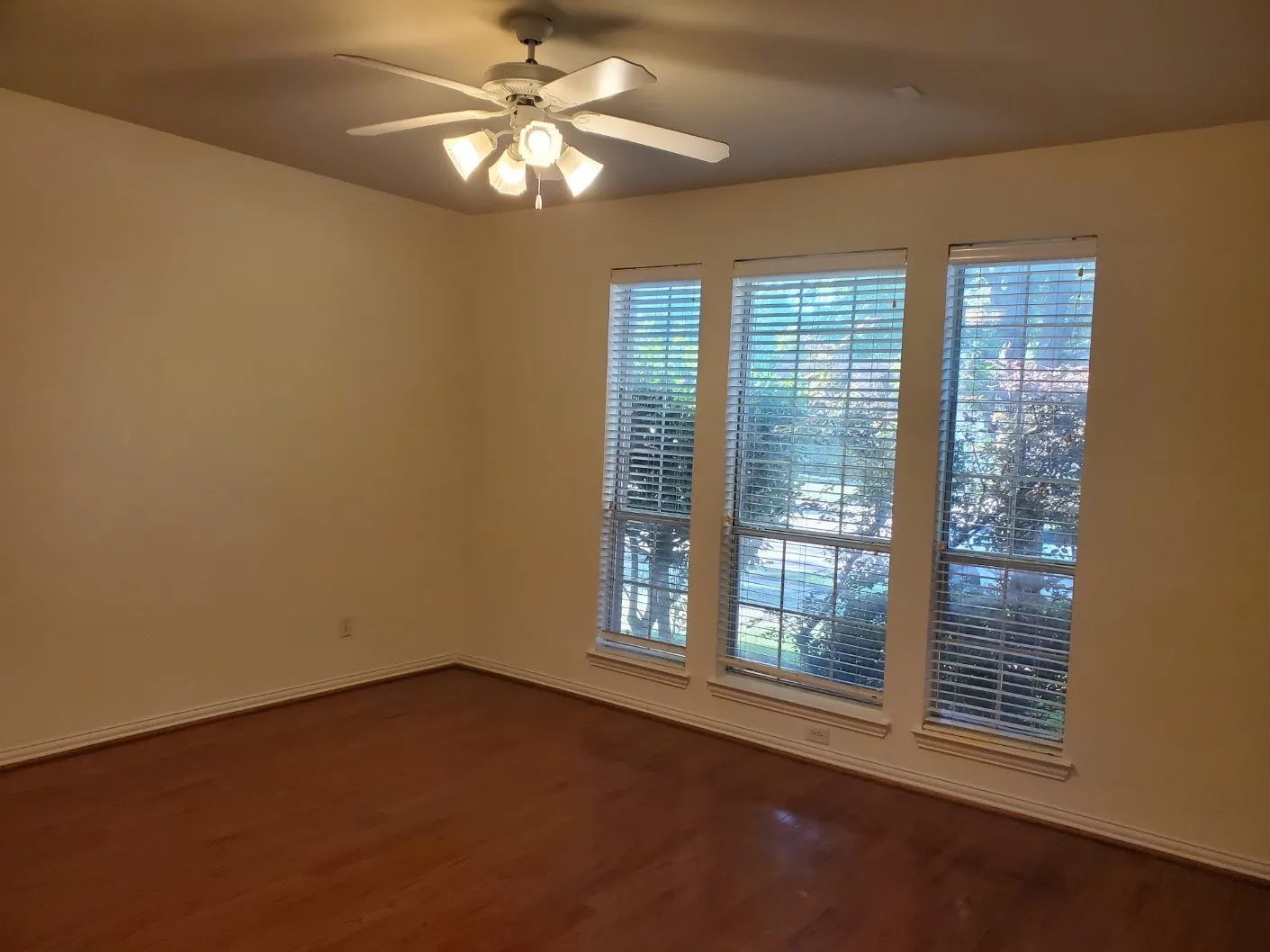 Expansive room featuring rich wood-finish flooring, three large windows with blinds, and a ceiling fan with integrated lighting