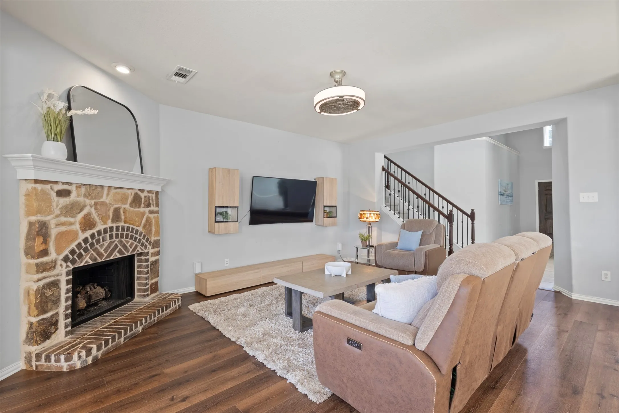 The living area features hardwood flooring, a stone fireplace with a brick hearth, and a staircase with dark wood handrails