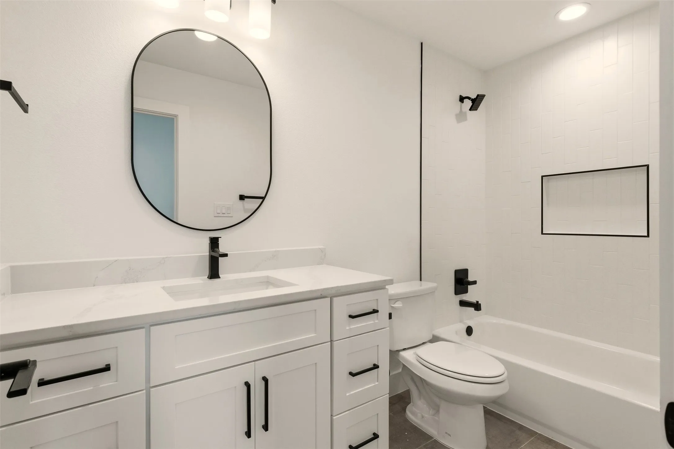 The bathroom features a modern vanity with a white countertop and dark-colored hardware, an oval mirror, and a tiled shower-bath combination with a built-in shelf
