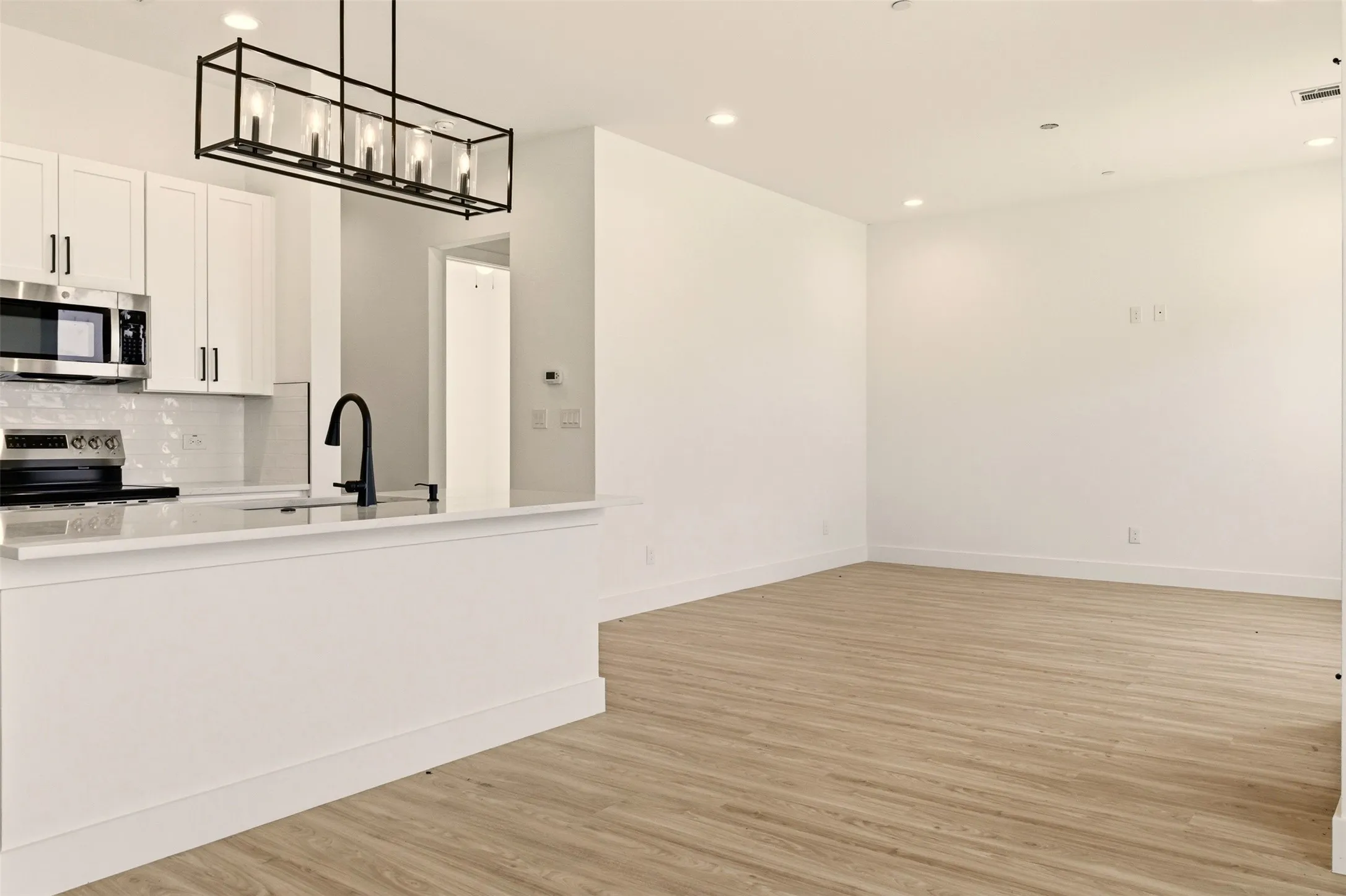 Open-concept living space with light wood-style flooring, recessed lighting, and a kitchen featuring white cabinetry, stainless steel appliances, and a modern black linear chandelier