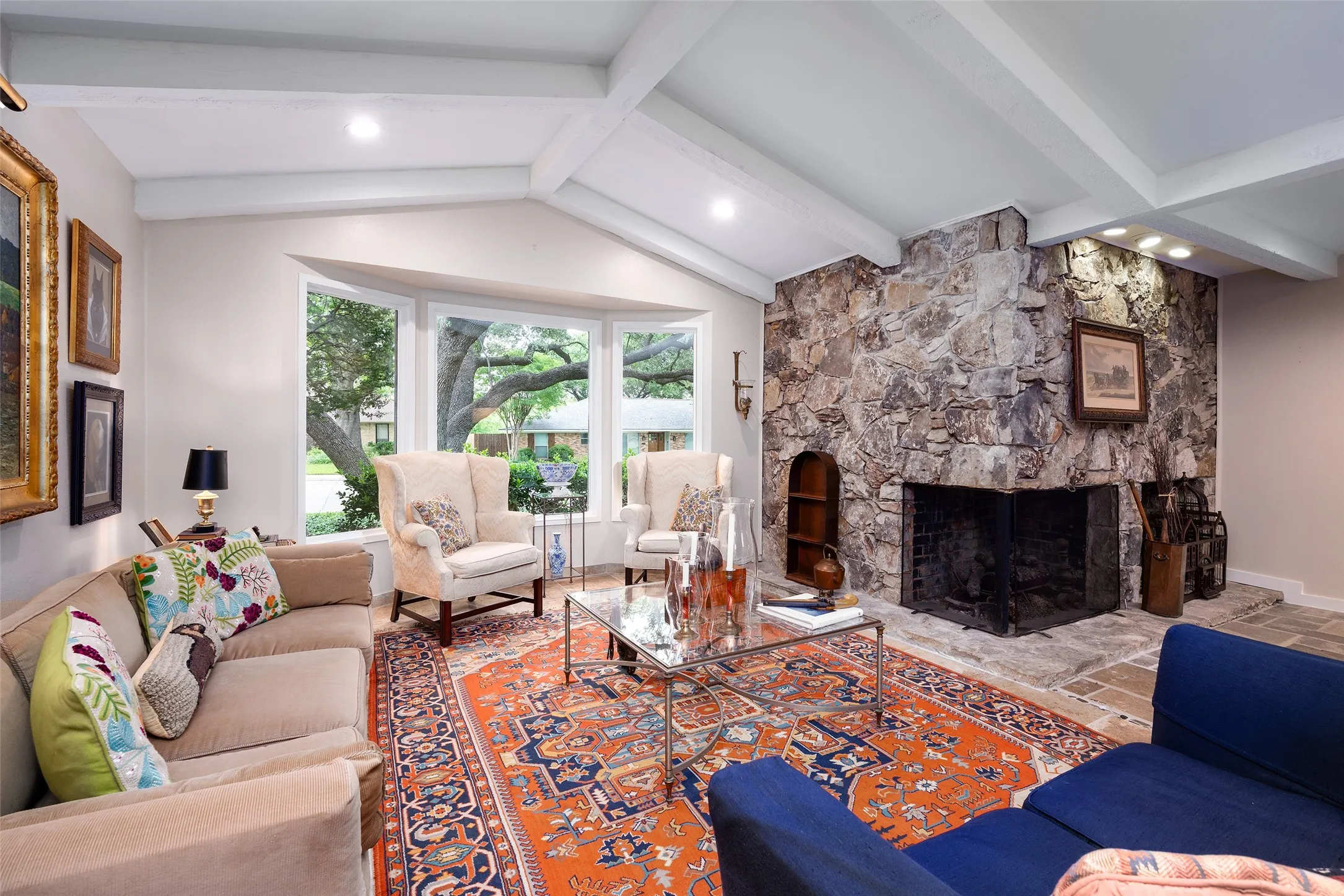 This living space features a vaulted ceiling with exposed beams, a natural stone fireplace, and a bay window providing views of the outdoors