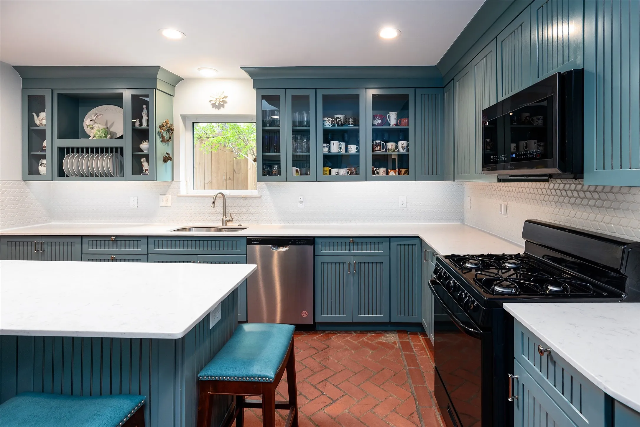The kitchen features blue cabinetry, white countertops, a white tile backsplash, a stainless steel dishwasher, and new black gas range.  Patterned brick flooring.