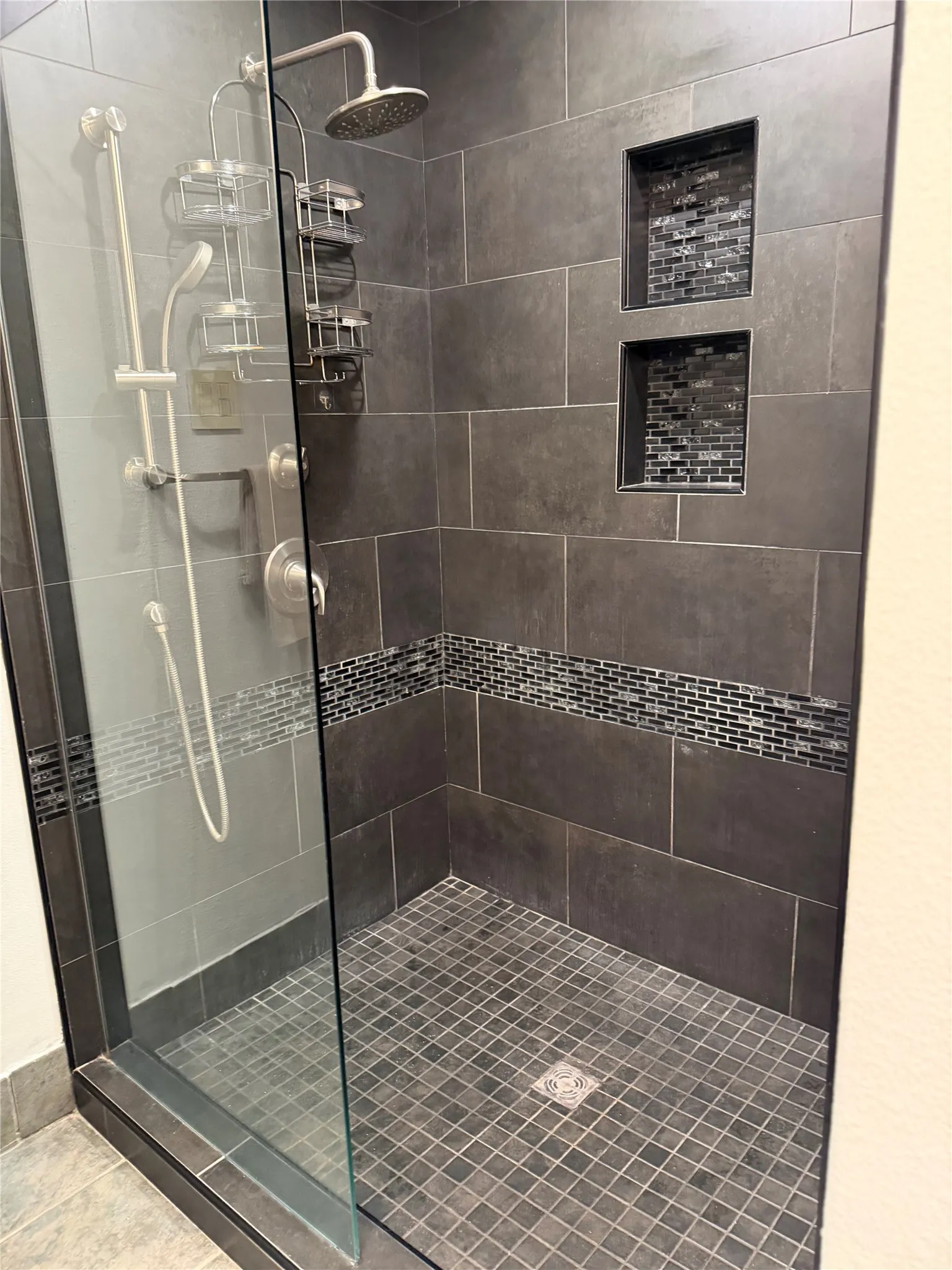 Enclosed shower with dark rectangular wall tiling, mosaic tile accents, recessed shelving, and a rain shower head