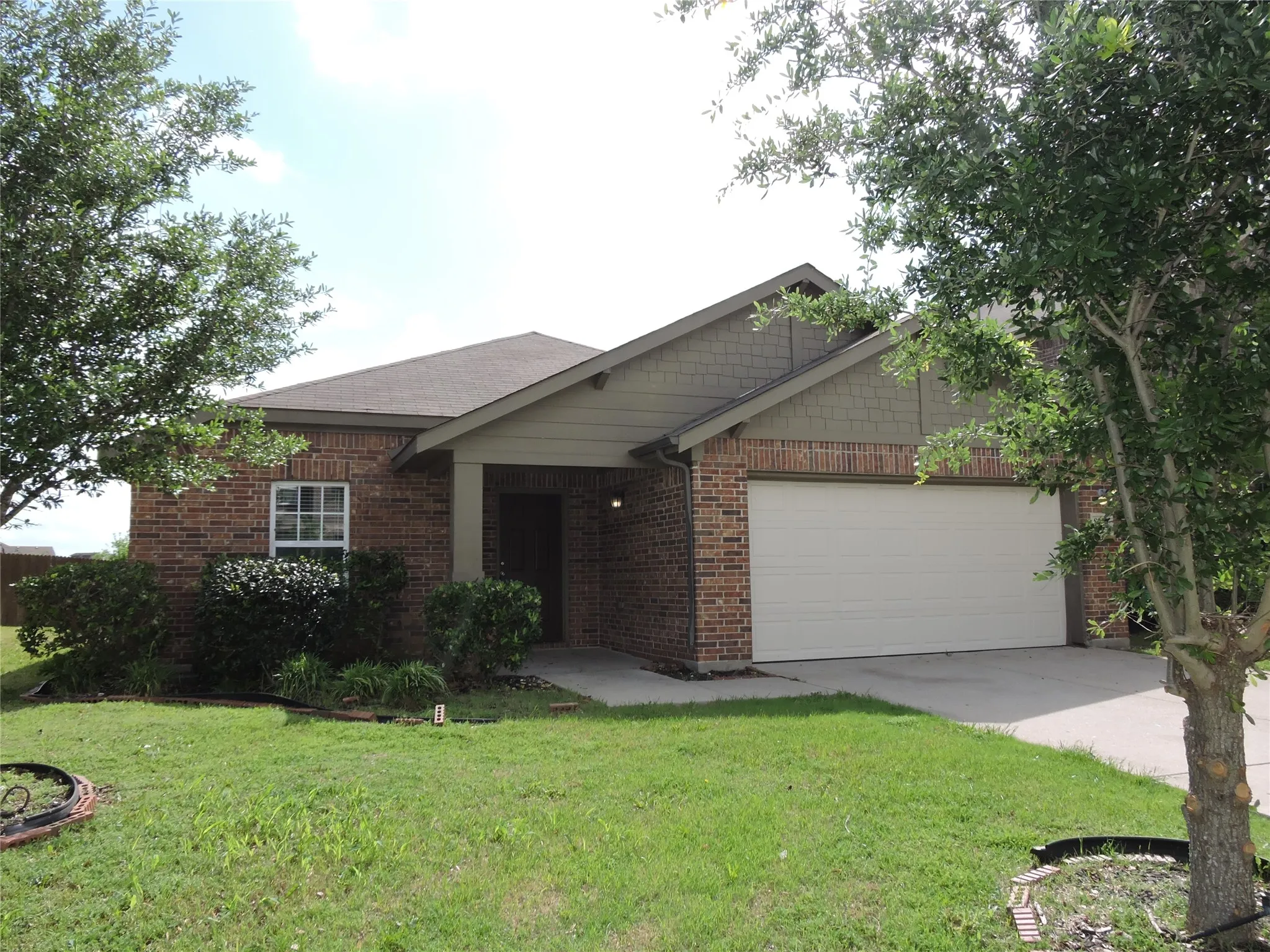 The property features a brick exterior with a front-facing garage, a well-maintained lawn, and established trees providing natural shade