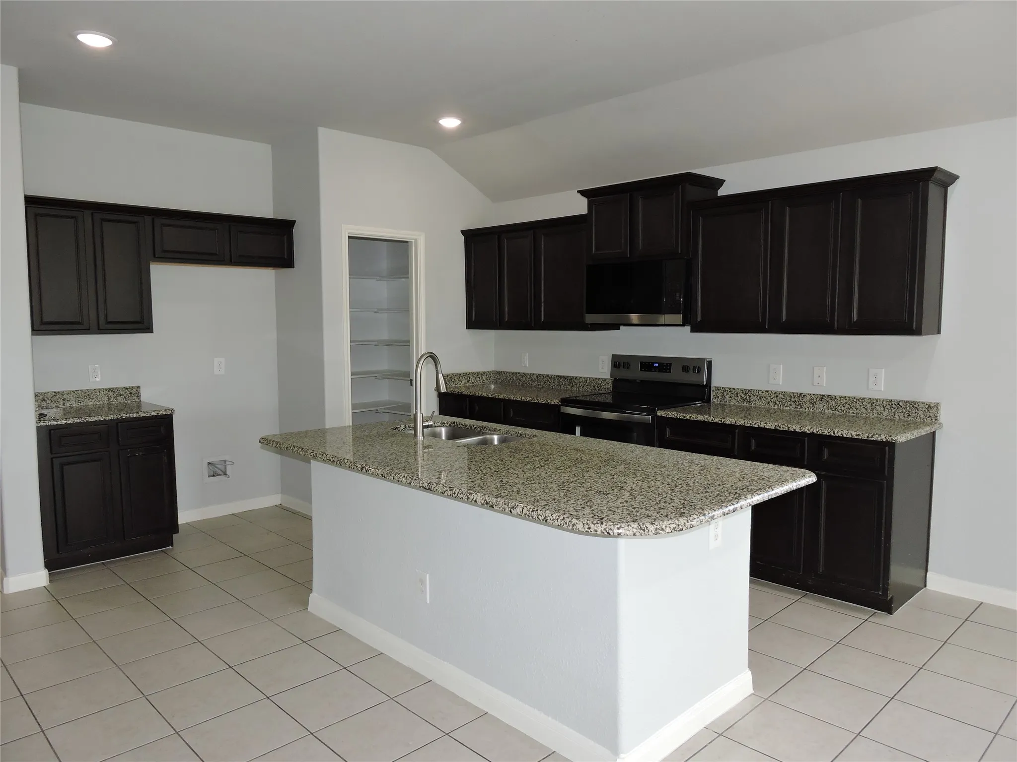 Kitchen featuring an island with a sink and a gooseneck faucet, dark cabinetry, light-colored granite countertops, and tile flooring