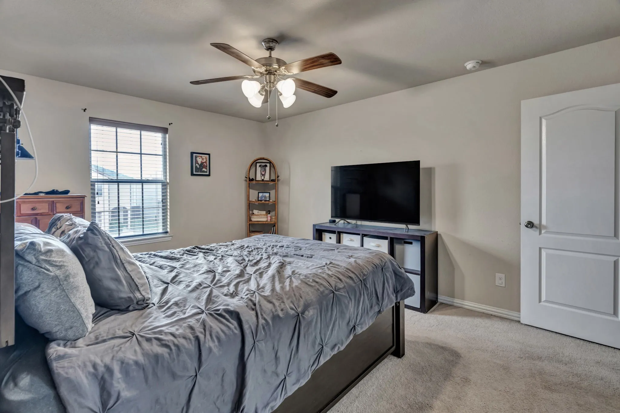 This bedroom features neutral-toned walls, carpet flooring, a window with blinds, and a ceiling fan with integrated lighting