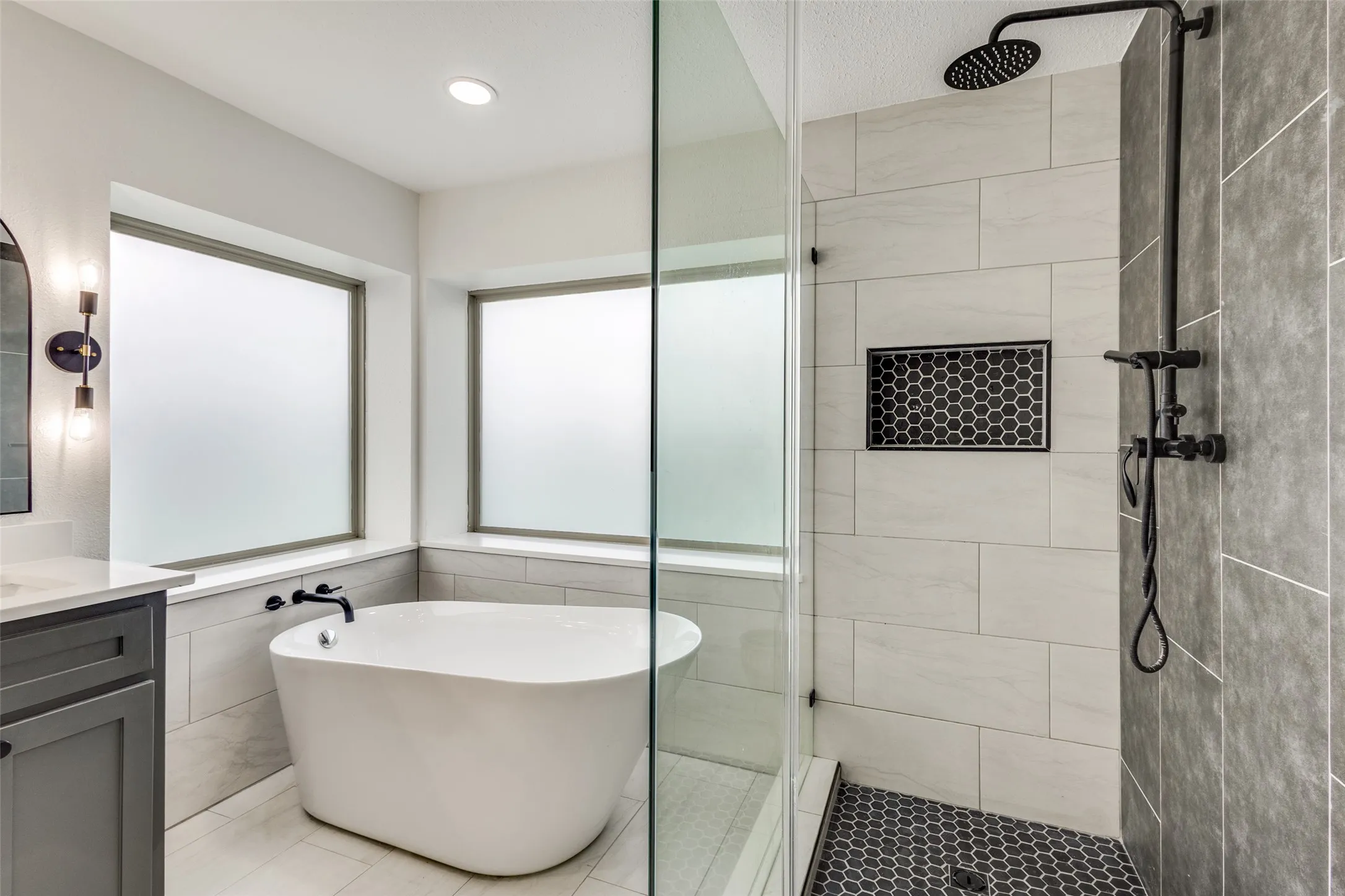 The bathroom features a freestanding soaking tub positioned beneath two windows, a walk-in shower with a glass enclosure, and a dark-toned vanity with a light countertop
