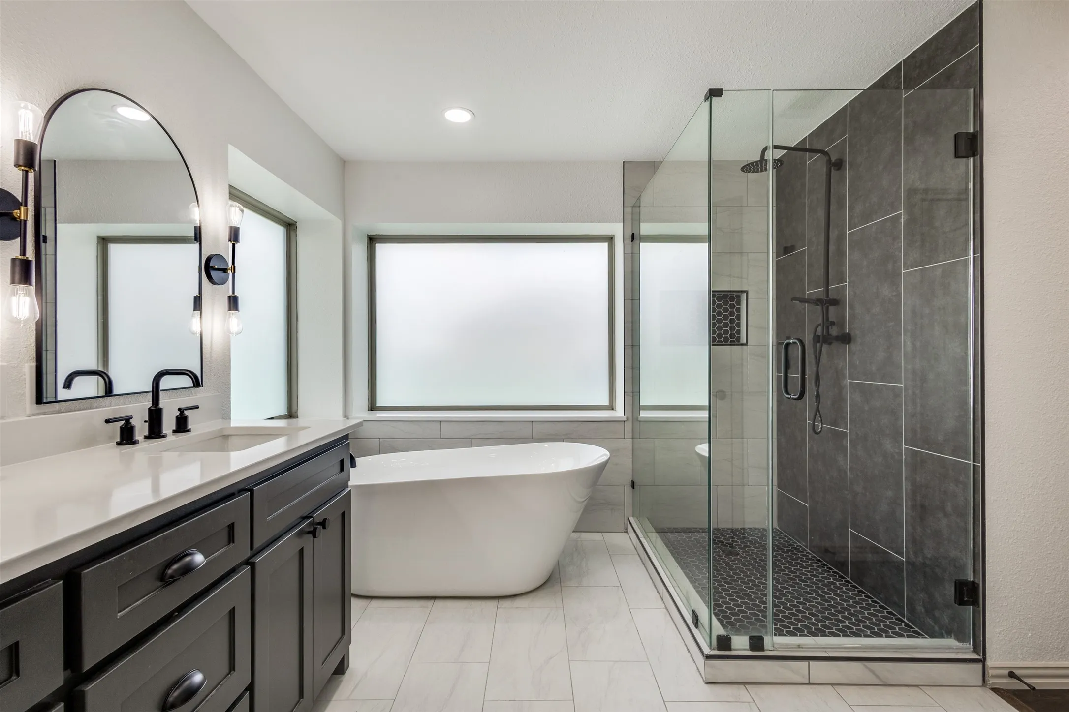Bathroom featuring a dark wood vanity with a white countertop, an arched mirror, a freestanding bathtub, and a glass-enclosed shower with dark tile