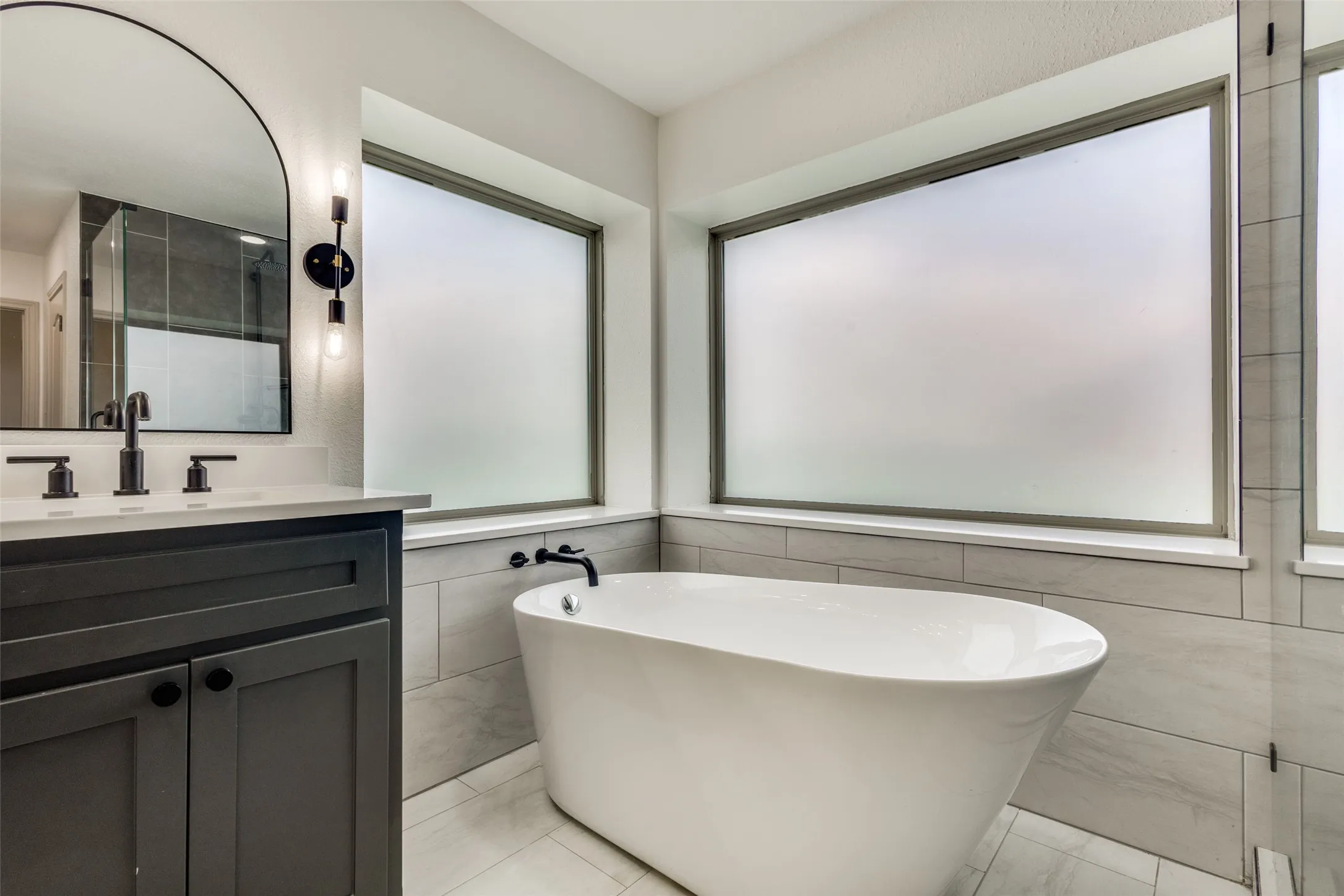Bathroom featuring a freestanding white bathtub, a dark-colored vanity with a white countertop and a black faucet, and a large arched mirror