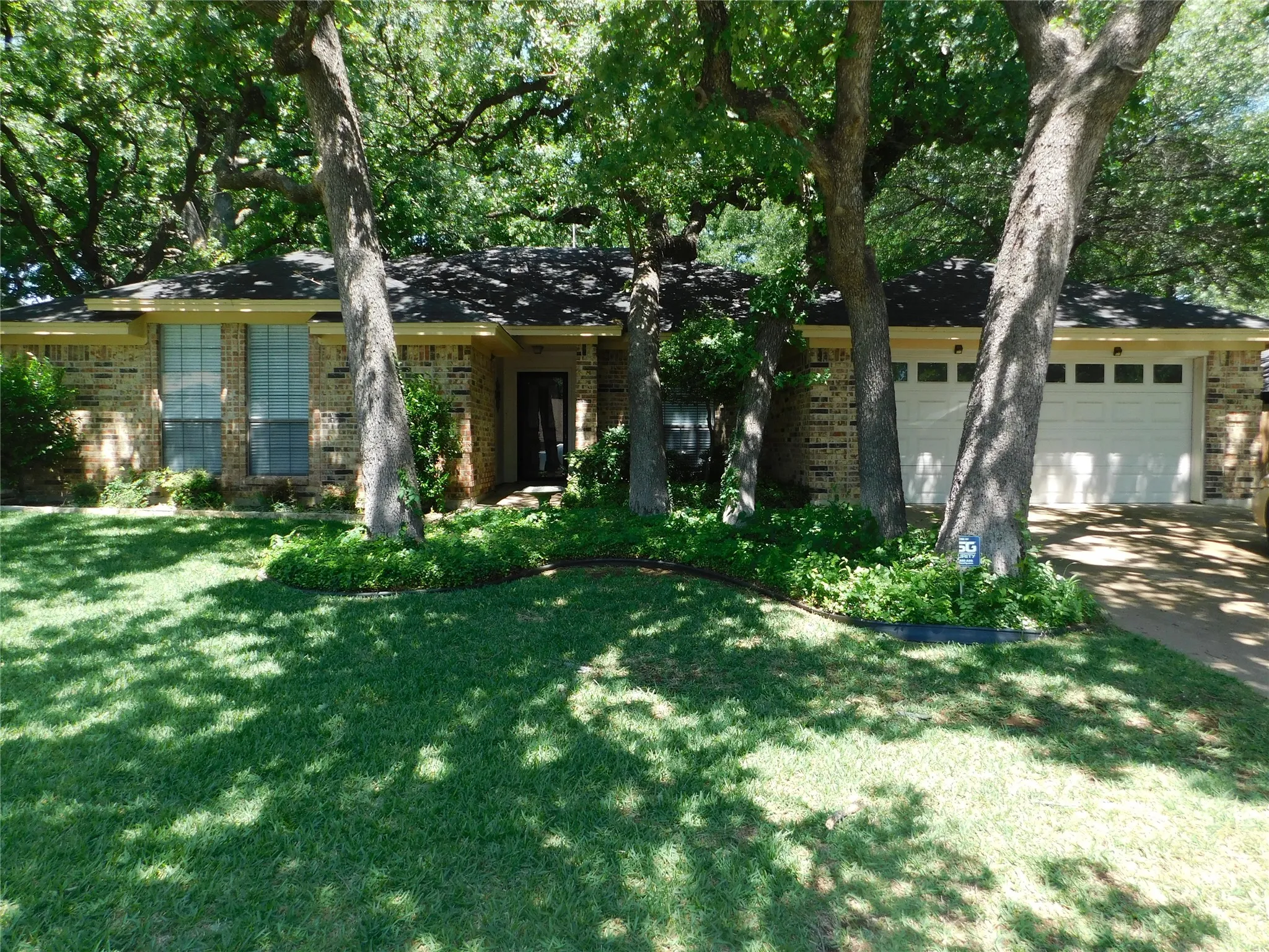 The property features a brick exterior, a well-maintained lawn, and mature trees providing natural shade