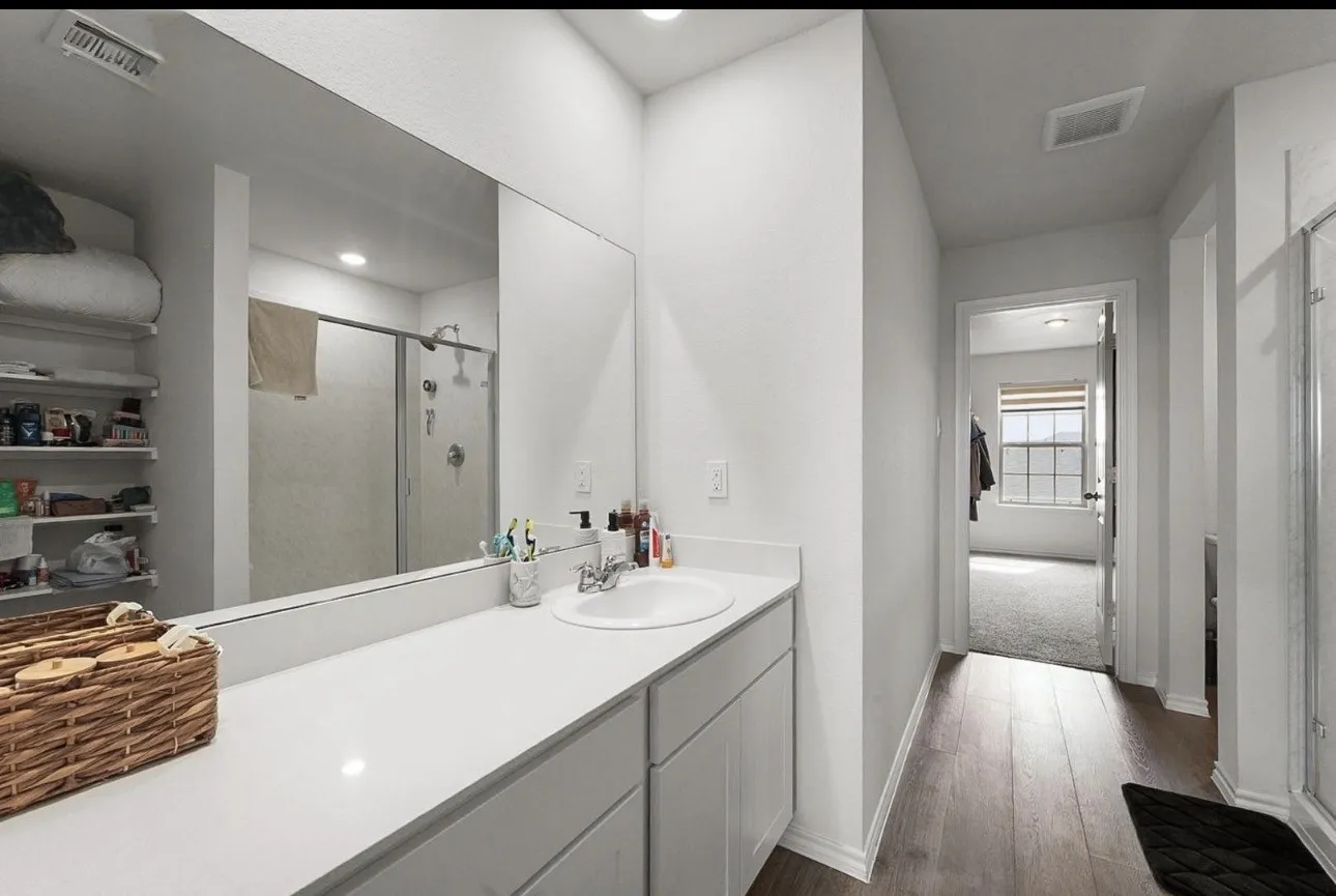 Spacious bathroom featuring a single vanity with an undermount sink, a large mirror, a glass-enclosed shower, built-in shelving, and wood-finish flooring