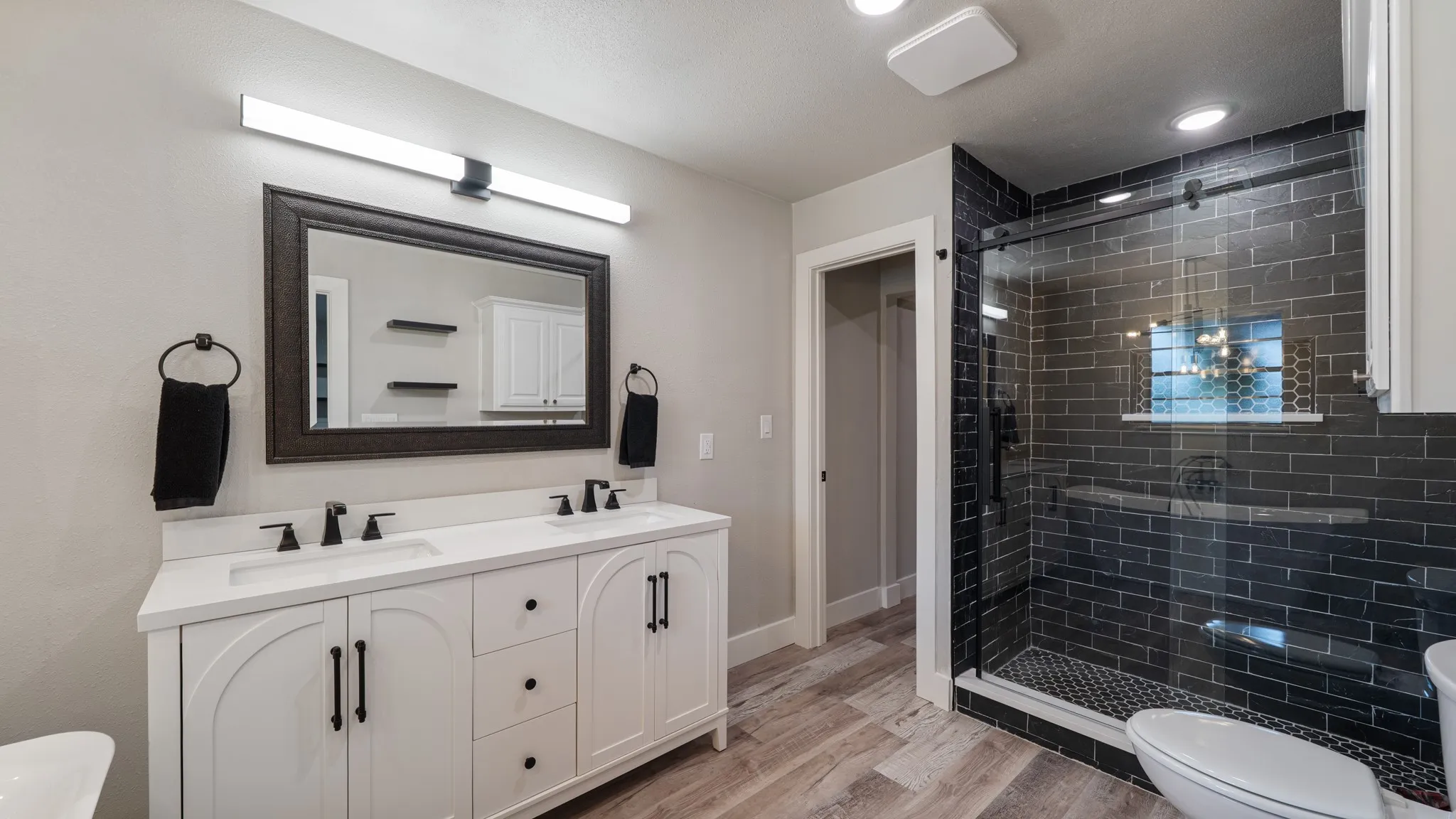 Bathroom featuring a double vanity with white cabinetry, a glass-enclosed shower with dark subway tiles, and wood-look flooring