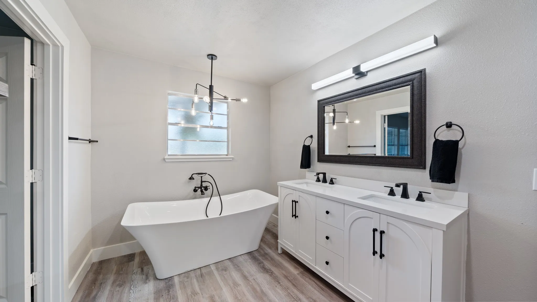 The bathroom features a freestanding tub, a window with privacy glass, a double vanity with white cabinetry and dark hardware, and a dark framed mirror with overhead lighting