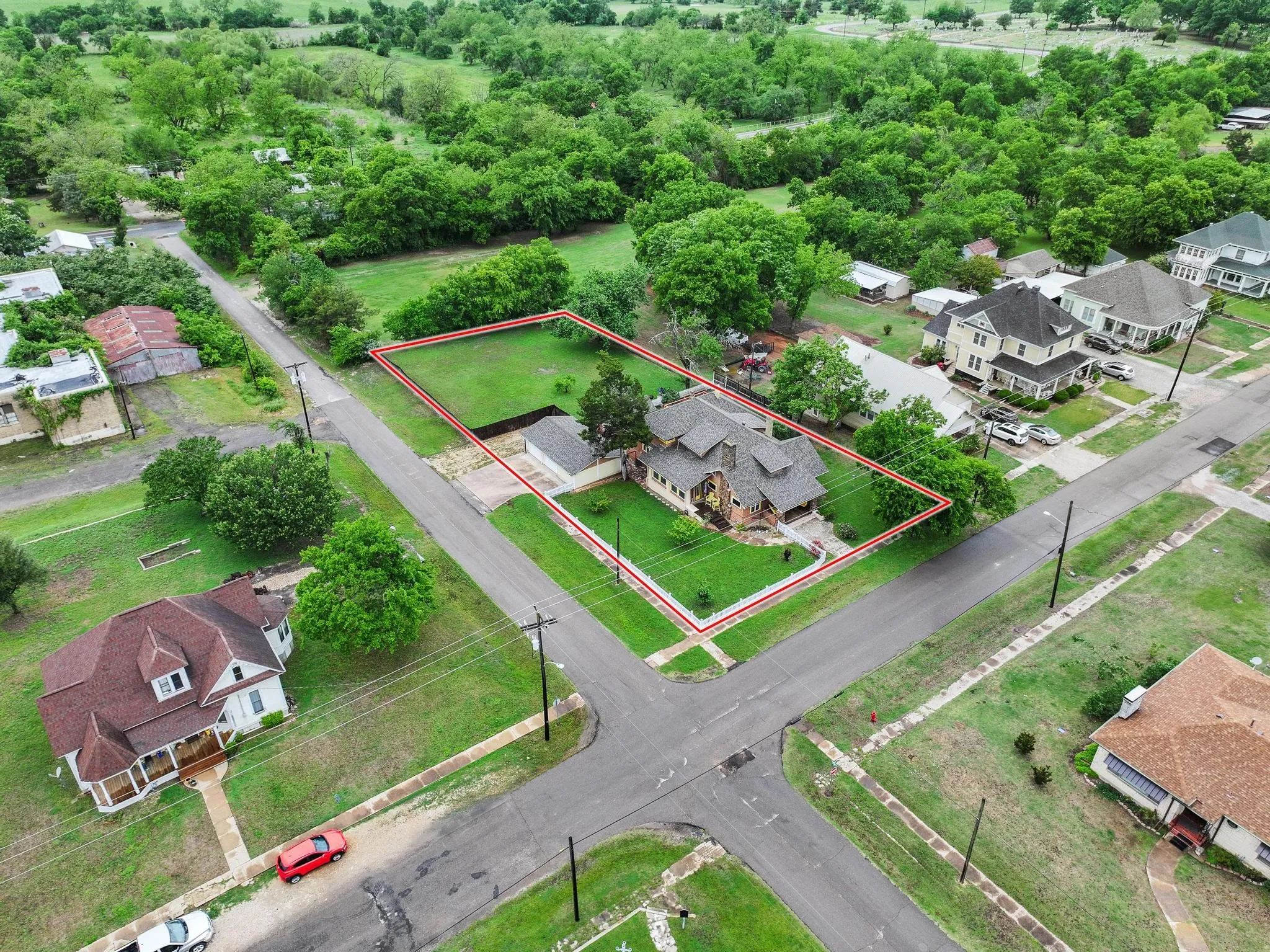 Aerial view showcasing the property's corner lot position, substantial green lawn, and mature trees, alongside a paved driveway leading to a garage