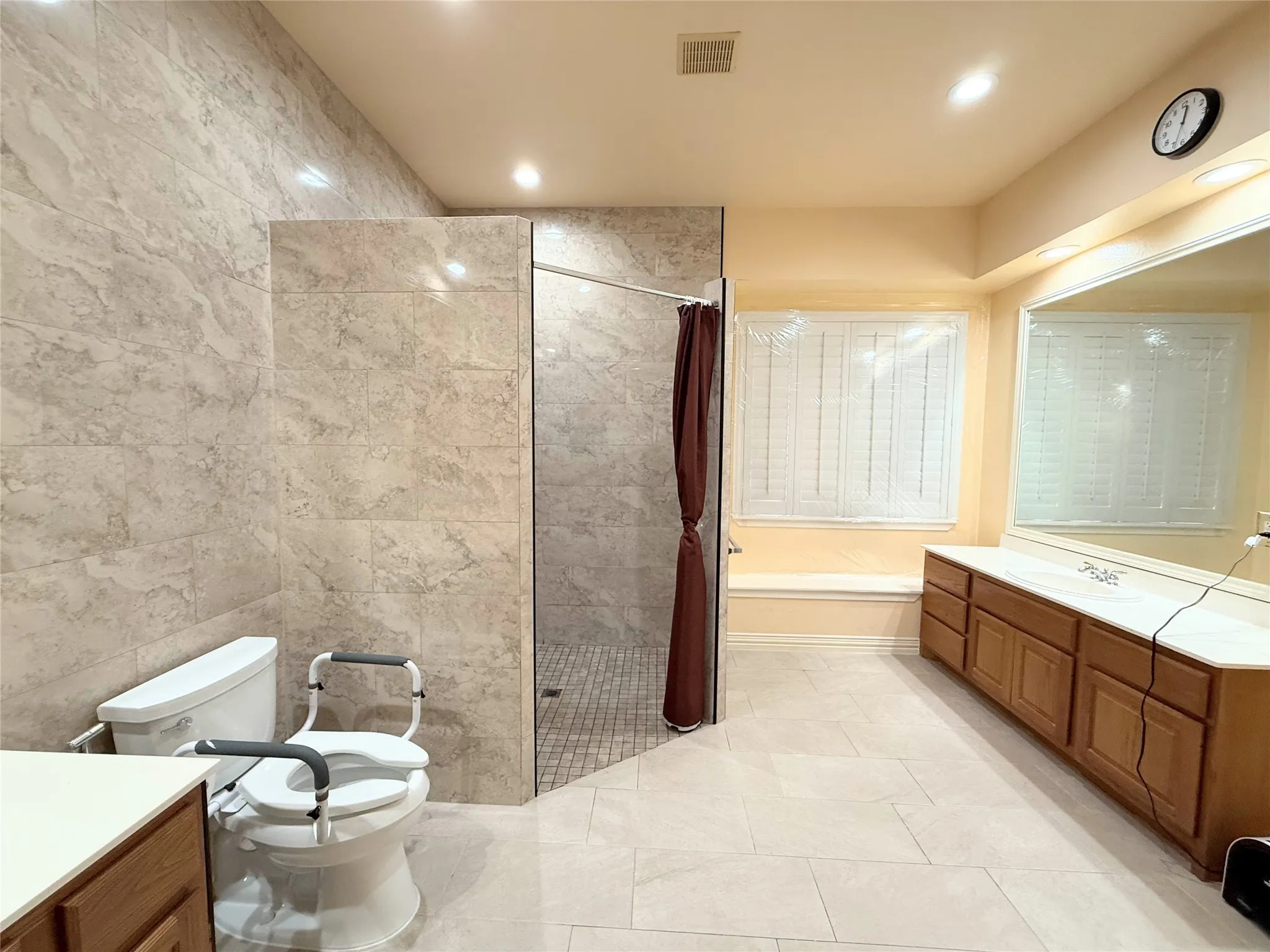 Spacious bathroom featuring a large walk-in shower with tiled walls, a double vanity with ample storage, a built-in window seat, and light-toned floor tiling