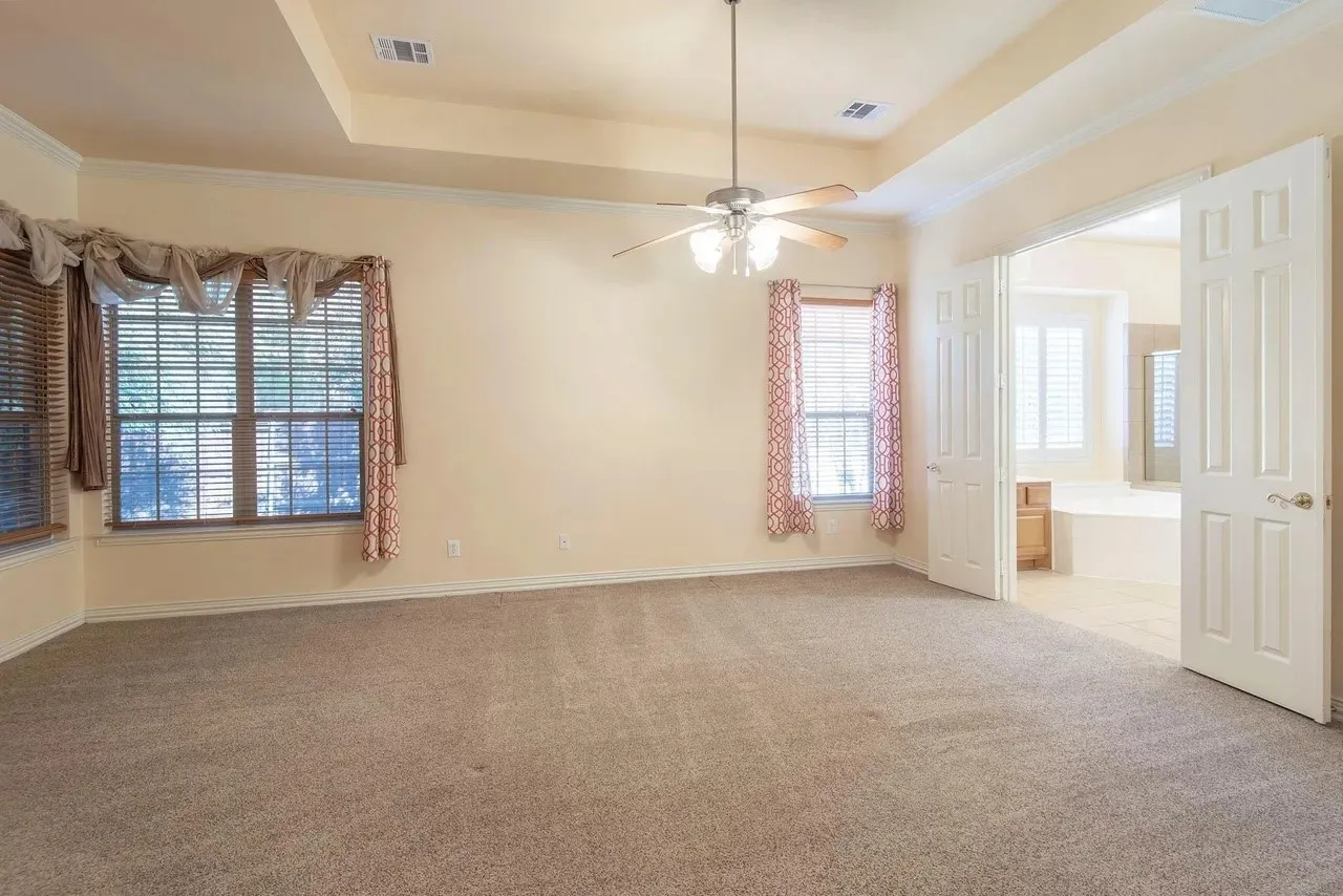 Spacious carpeted room featuring a tray ceiling with a ceiling fan, multiple windows with blinds, and double doors opening to an adjoining space with a built-in tub