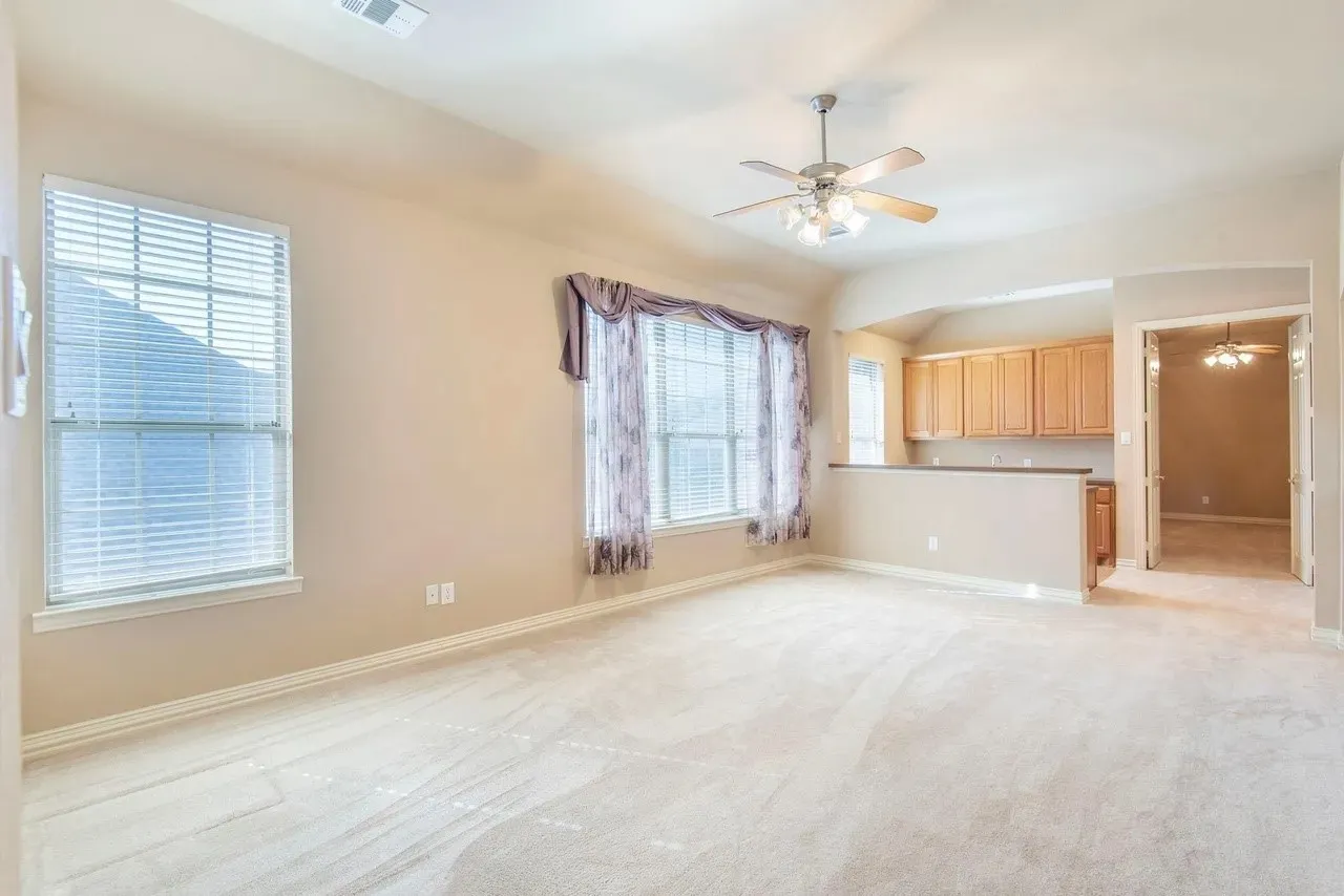 Open-concept living area featuring light-toned carpeting, multiple windows with blinds, a ceiling fan with integrated lighting, and a breakfast bar with built-in cabinetry