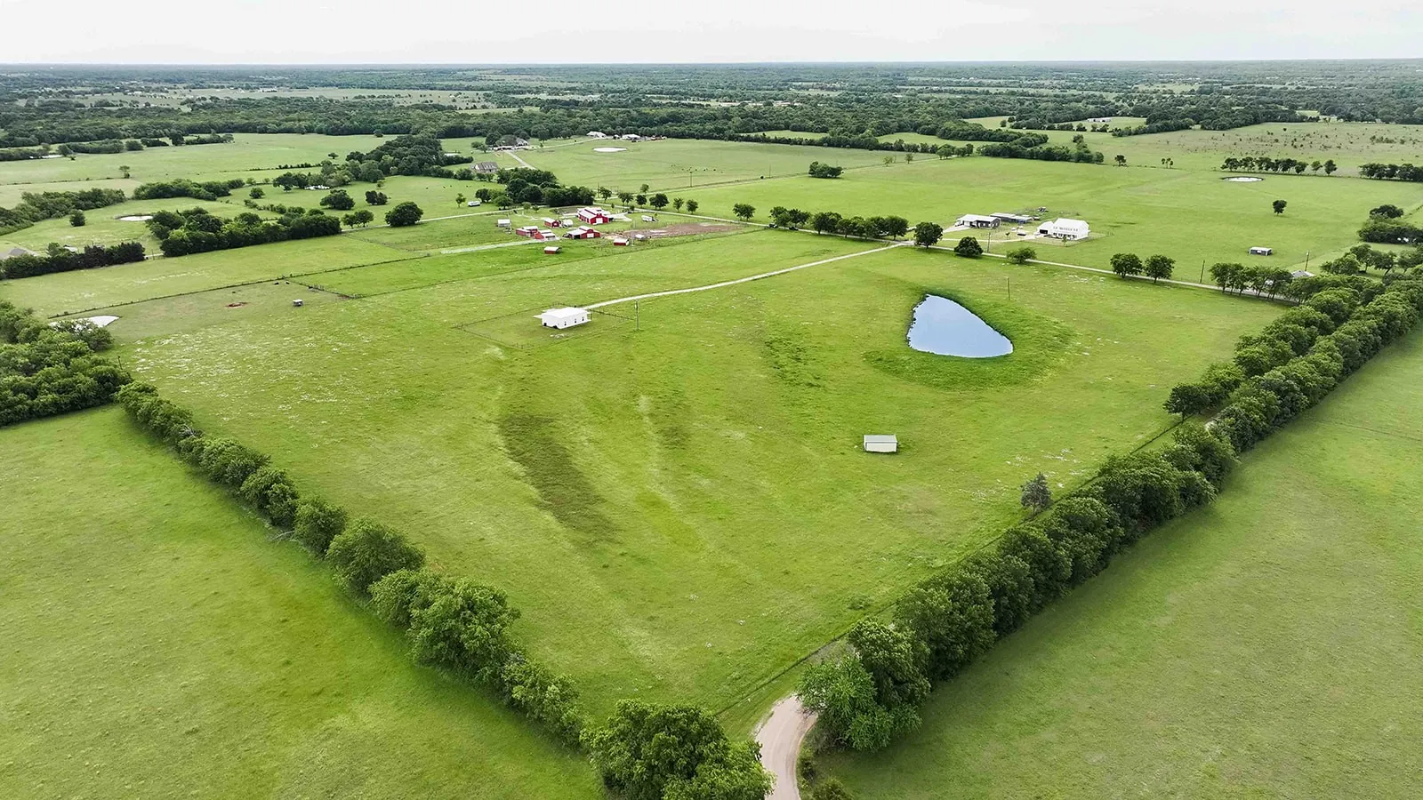 Expansive green acreage featuring a private pond, perimeter tree lines, and multiple structures