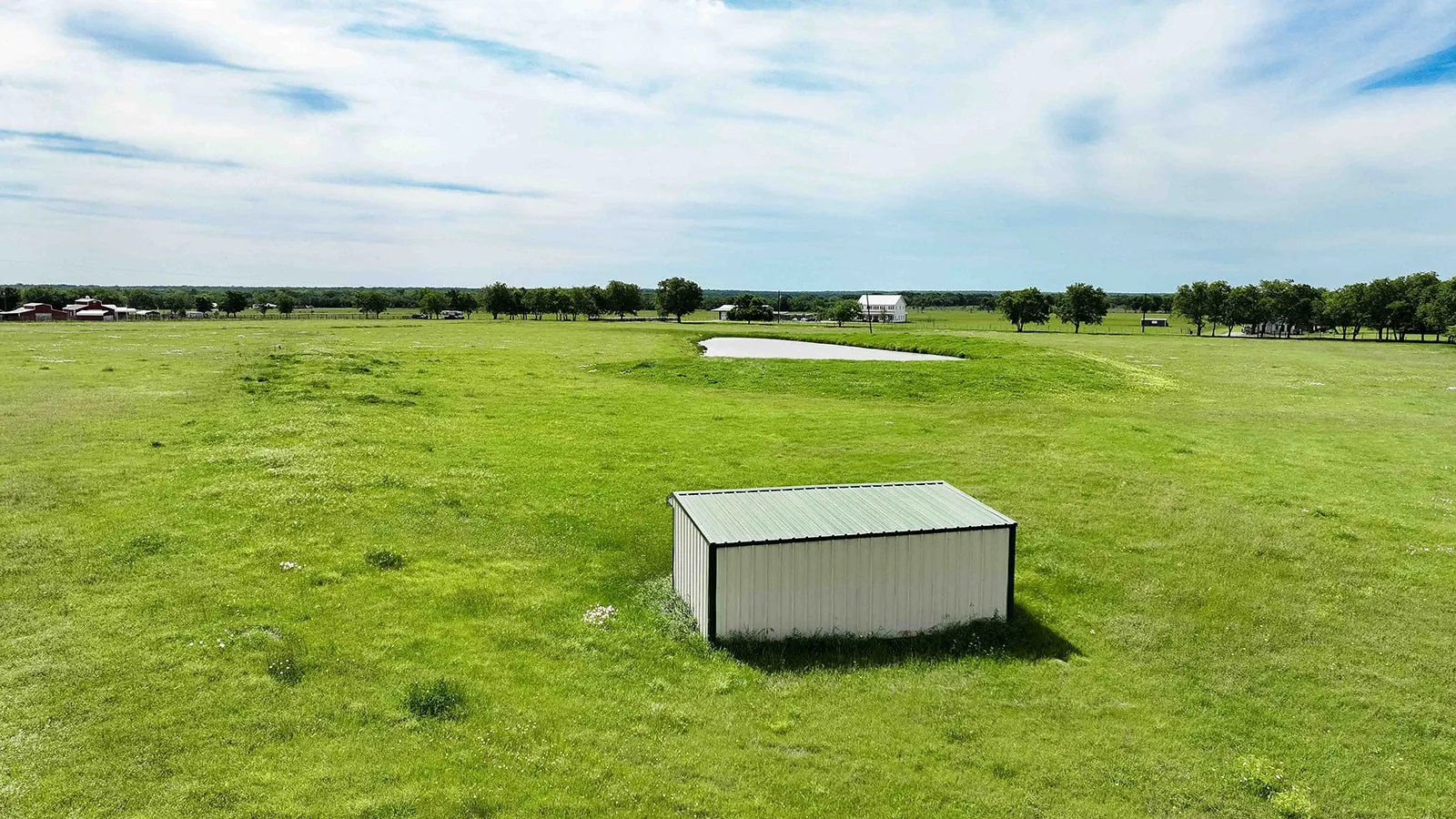 Expansive green pasture with a white metal storage shed featuring a green roof, a visible pond, and a distant white structure