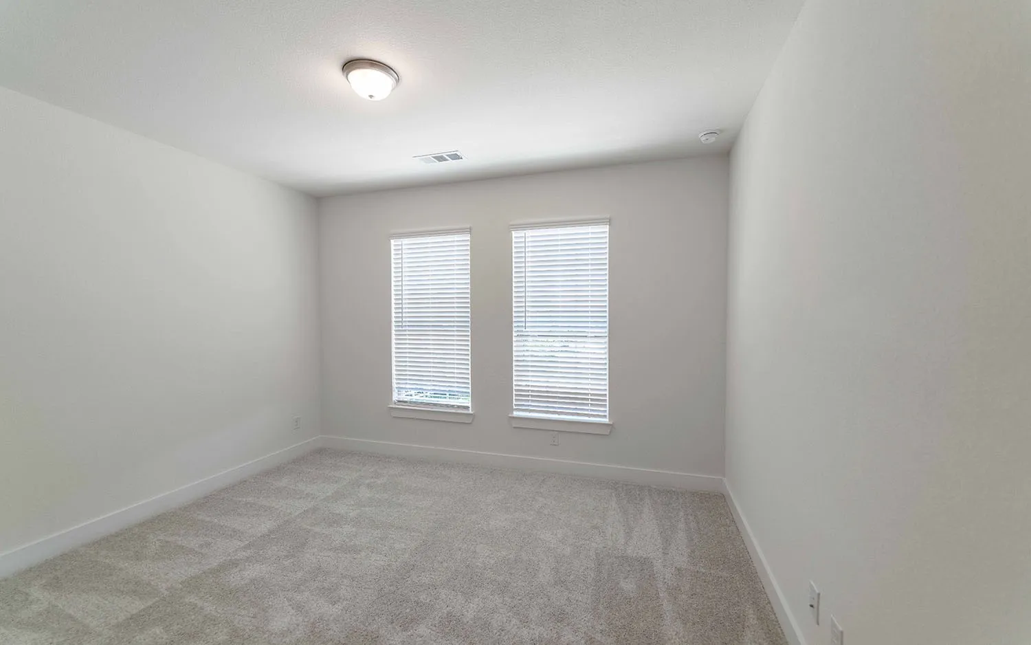Neutral tone walls featuring two windows with horizontal blinds, overhead flush mount lighting, and grey patterned carpeting