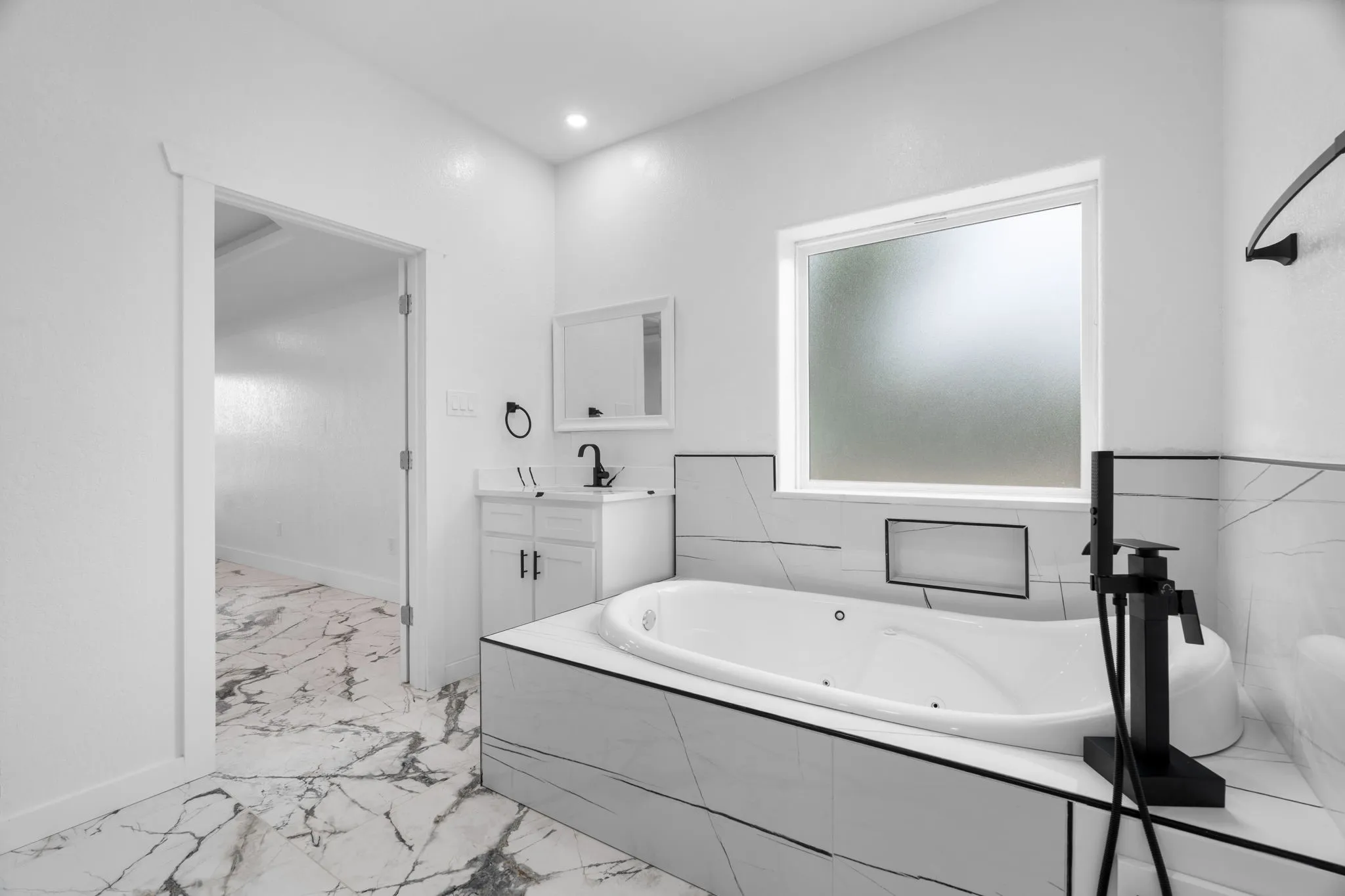 Bathroom featuring a built-in jetted tub with matte black fixtures, marble-patterned tile surround, and a frosted window