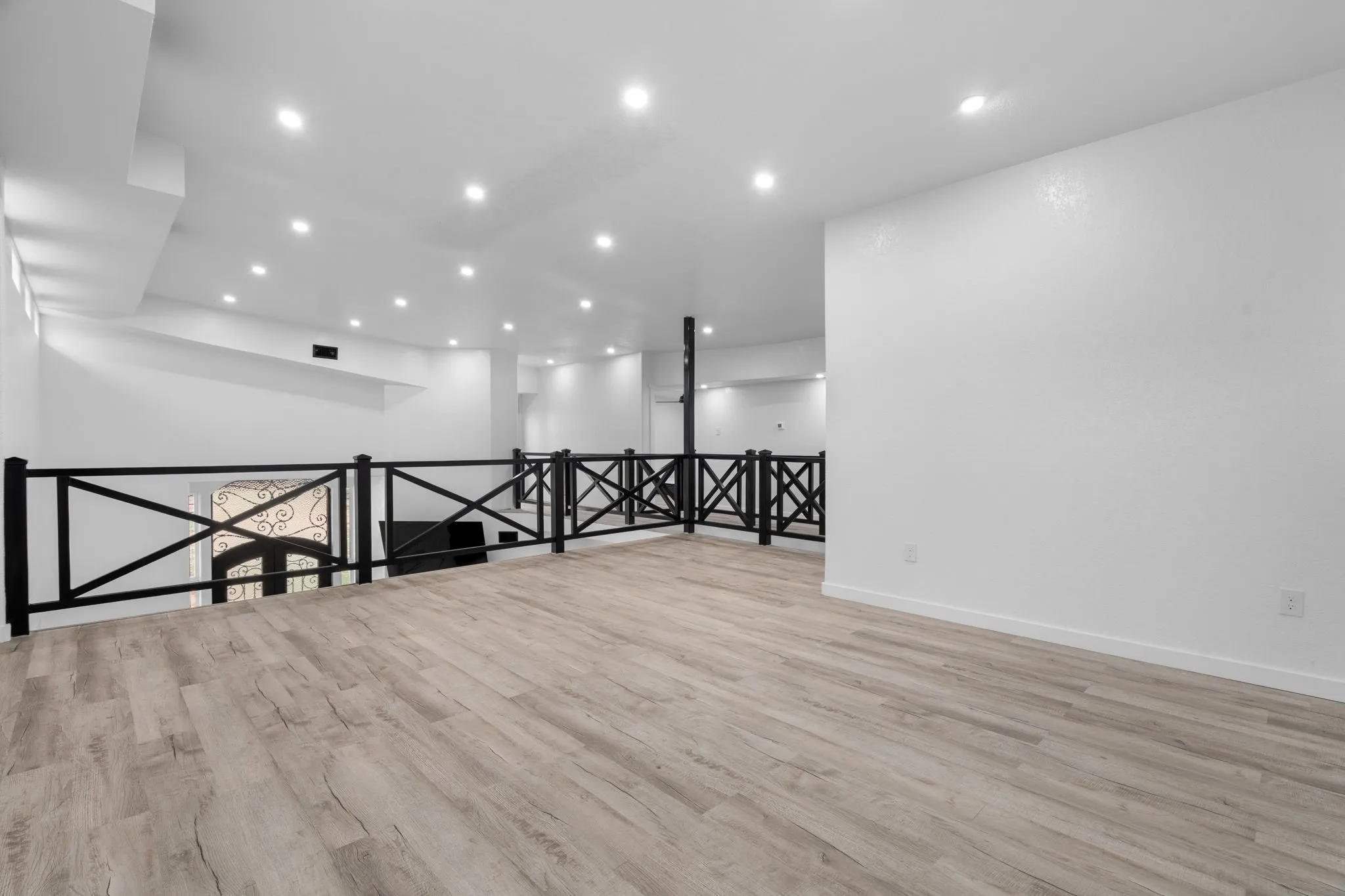 Spacious loft area featuring wood-finish flooring, white walls, recessed lighting, and a black railing with a decorative X-pattern