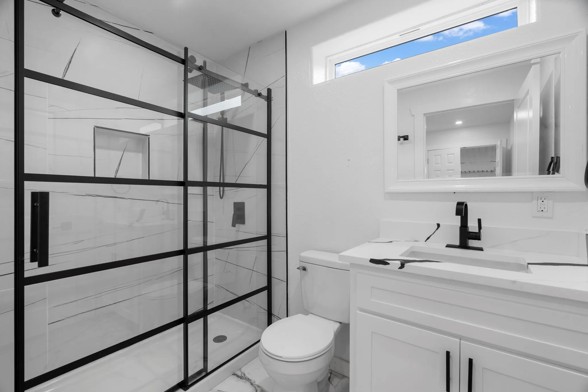 Modern bathroom featuring a glass-enclosed shower with black grid accents, white marble-look wall tiling, a white vanity with a rectangular undermount sink, and a black faucet